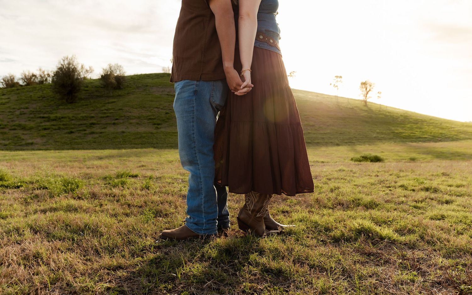 Couple holding hands in a sunlit green field, woman in brown skirt and boots, man in jeans, romantic outdoor setting.