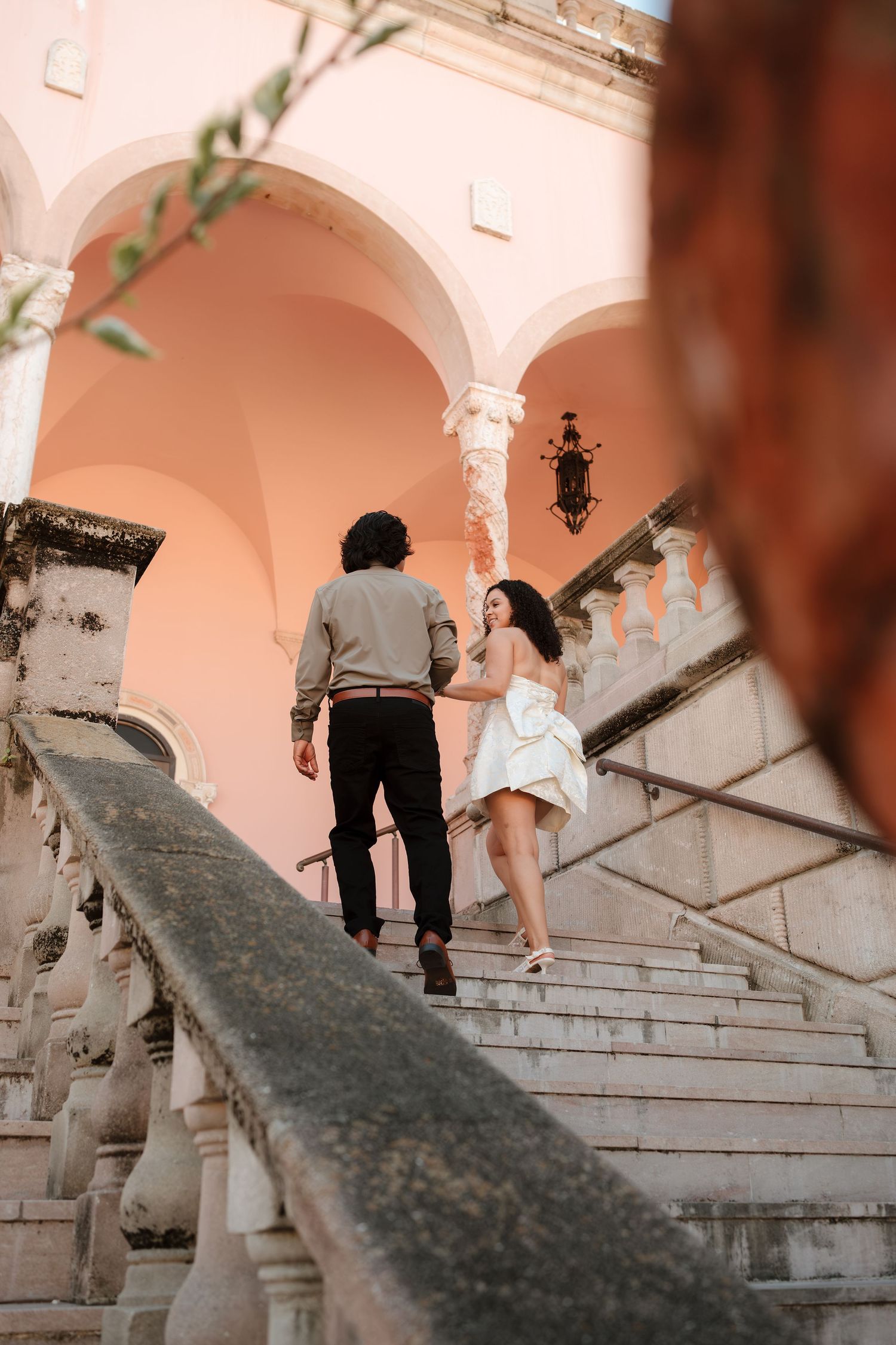 Couple in wedding attire holding hands while ascending ornate stone stairs of a pink historic building.