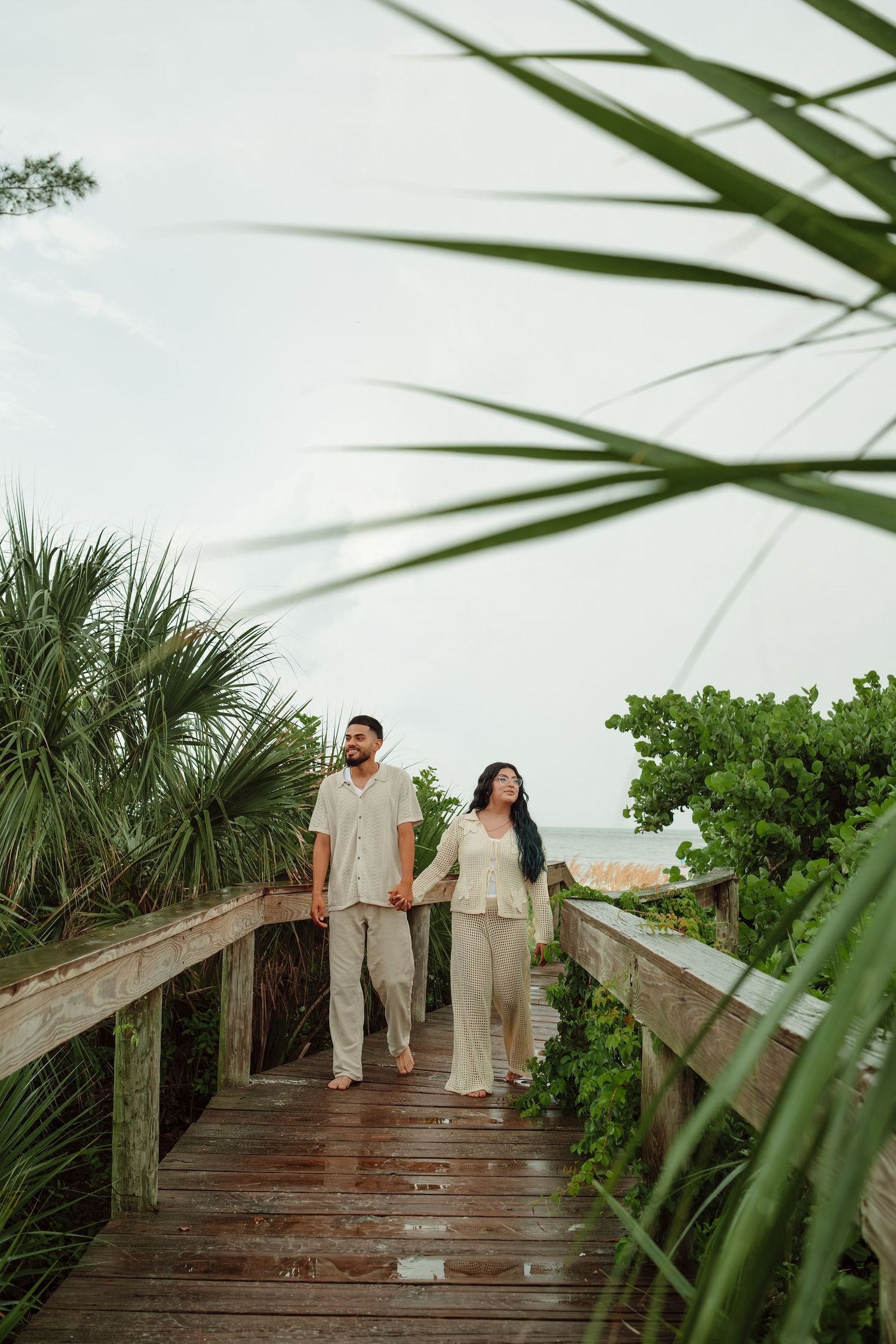 Couple in matching beige outfits holding hands on a wooden boardwalk surrounded by tropical greenery near the beach.