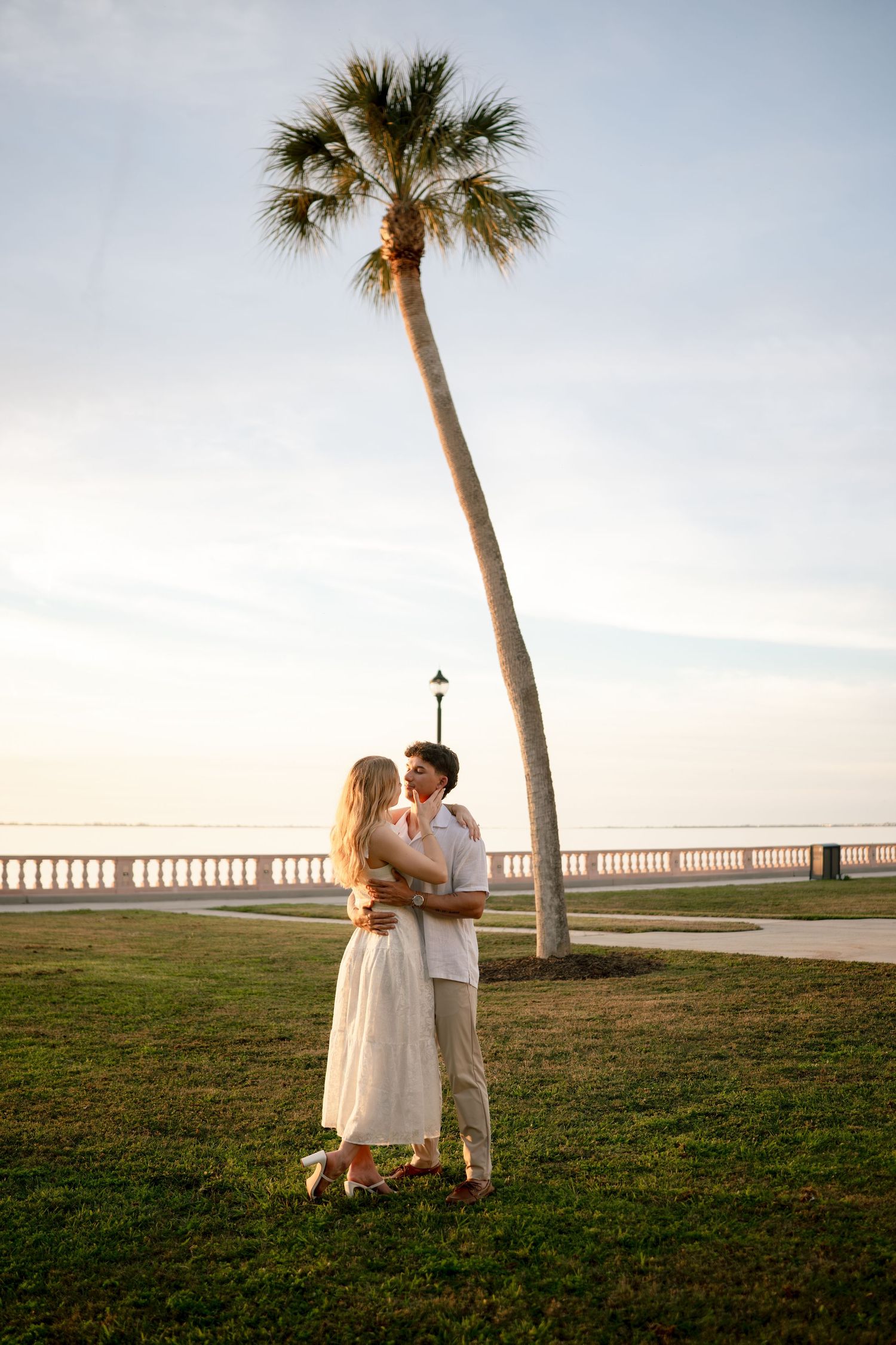 Couple embracing near a tall palm tree at sunset by a waterfront promenade, dressed in white and beige outfits.