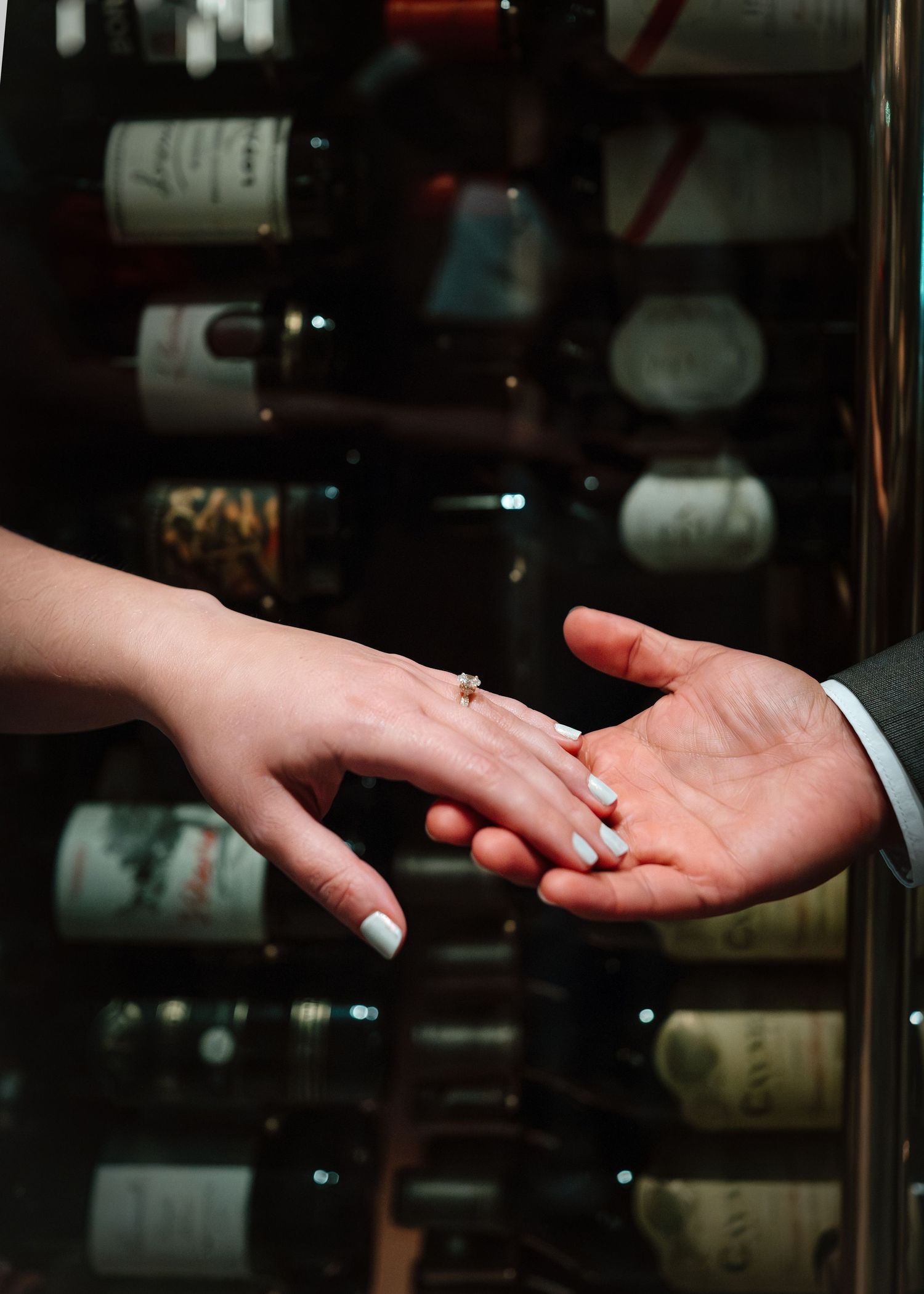 Two hands touching near a wine rack, with an engagement ring visible on the woman's finger.