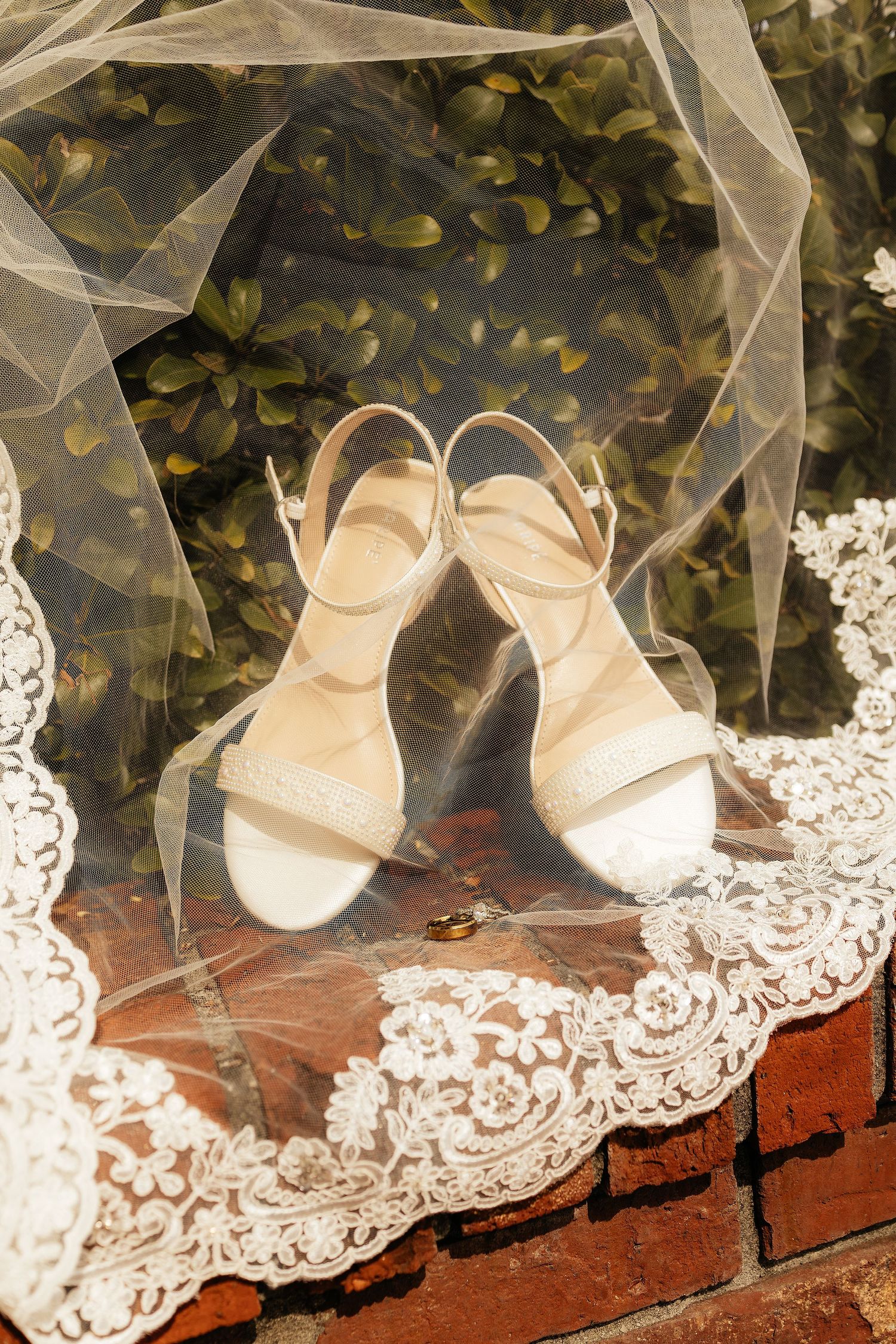 Bridal heels and lace veil displayed on brick surface with greenery backdrop.