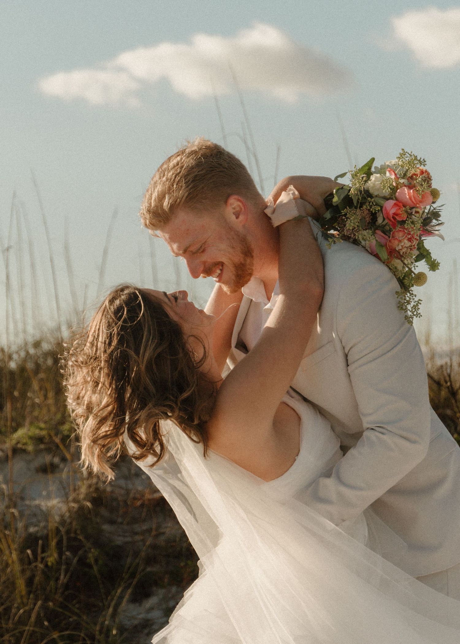 A groom lifts his bride in a wedding bouquet moment on a beach, both laughing joyfully under a blue sky.