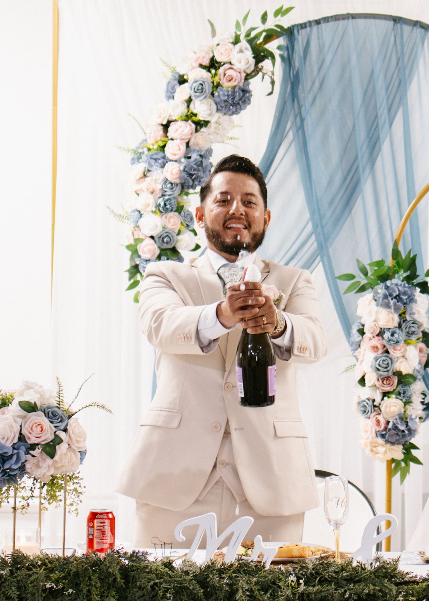 Groom in beige suit opens champagne bottle at wedding reception with floral arch backdrop.