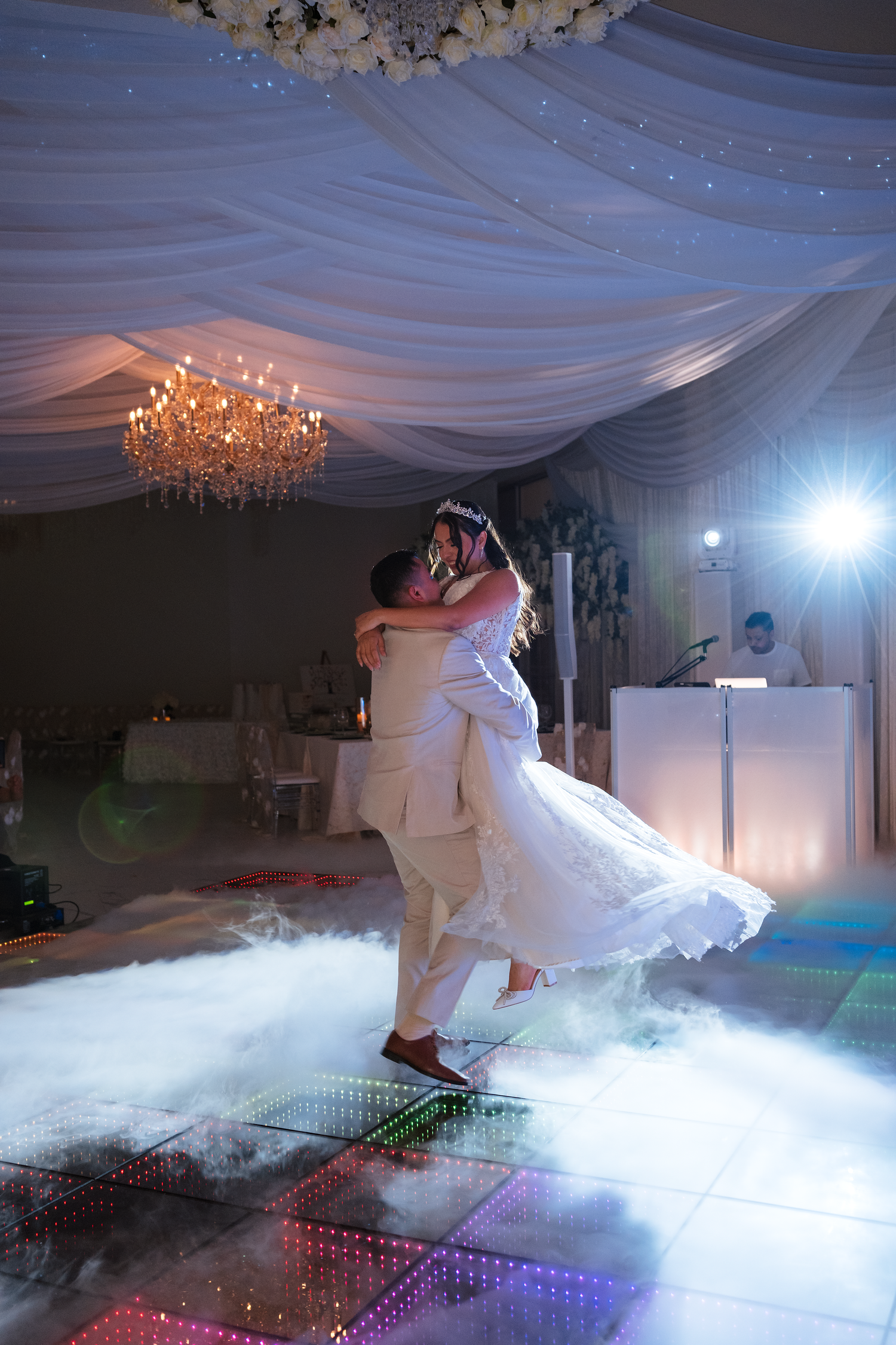 Groom lifts bride during romantic first dance on glowing dance floor with chandelier above.