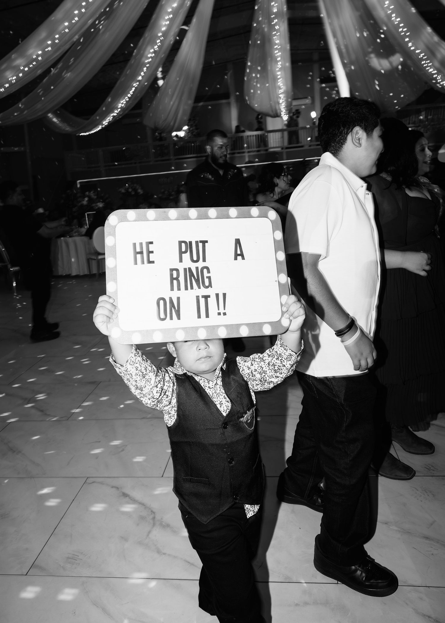 Child holds 'He Put A Ring On It' sign at wedding reception in black and white photo.