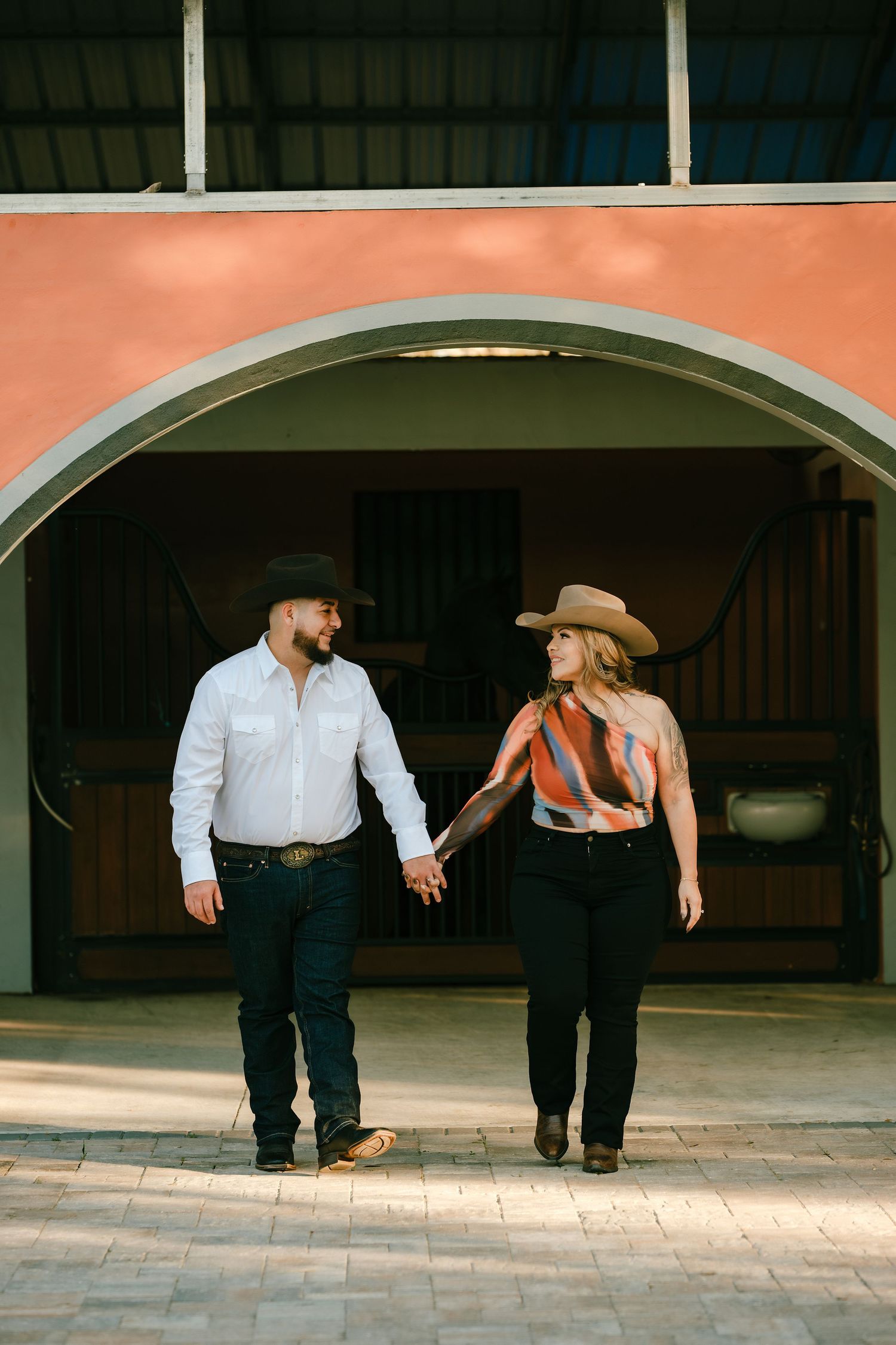 Couple in cowboy hats holding hands walking outside a rustic stable with an arched entrance.