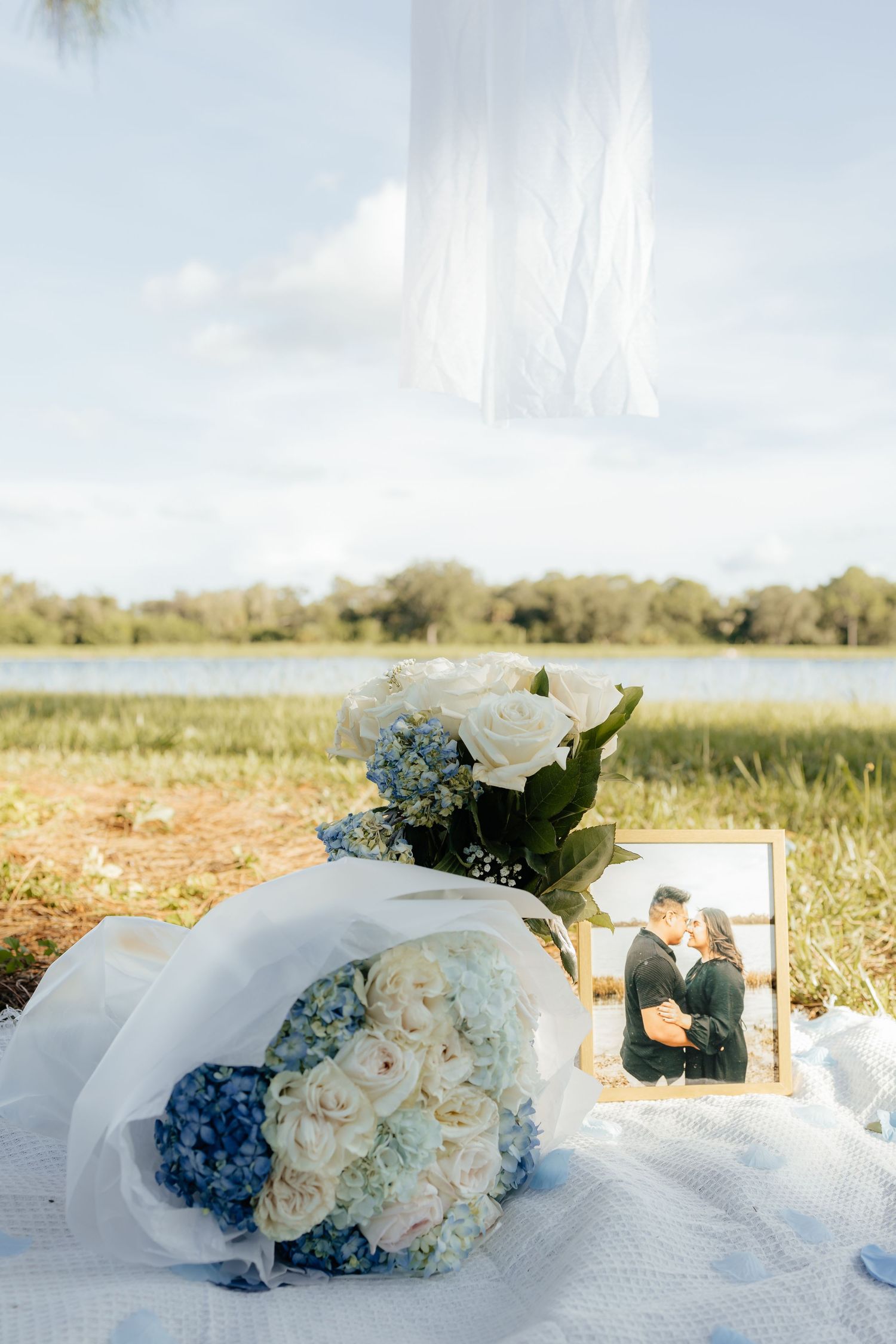 White roses and blue hydrangea wedding bouquet beside a couple's photo on a white blanket near a serene lakeside backdrop.