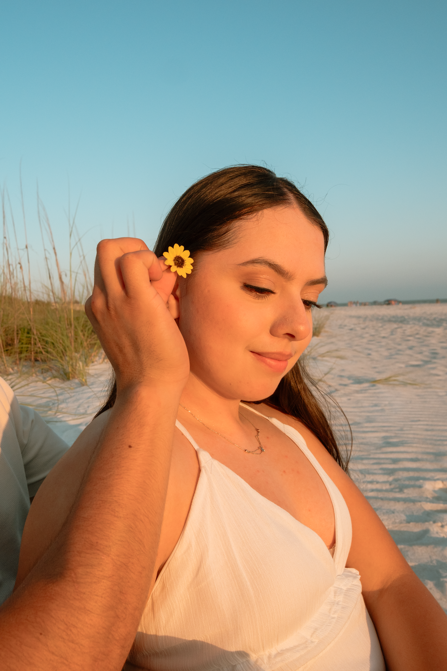 A young woman in a white dress smiles as a sunflower is tucked behind her ear on a sandy beach.