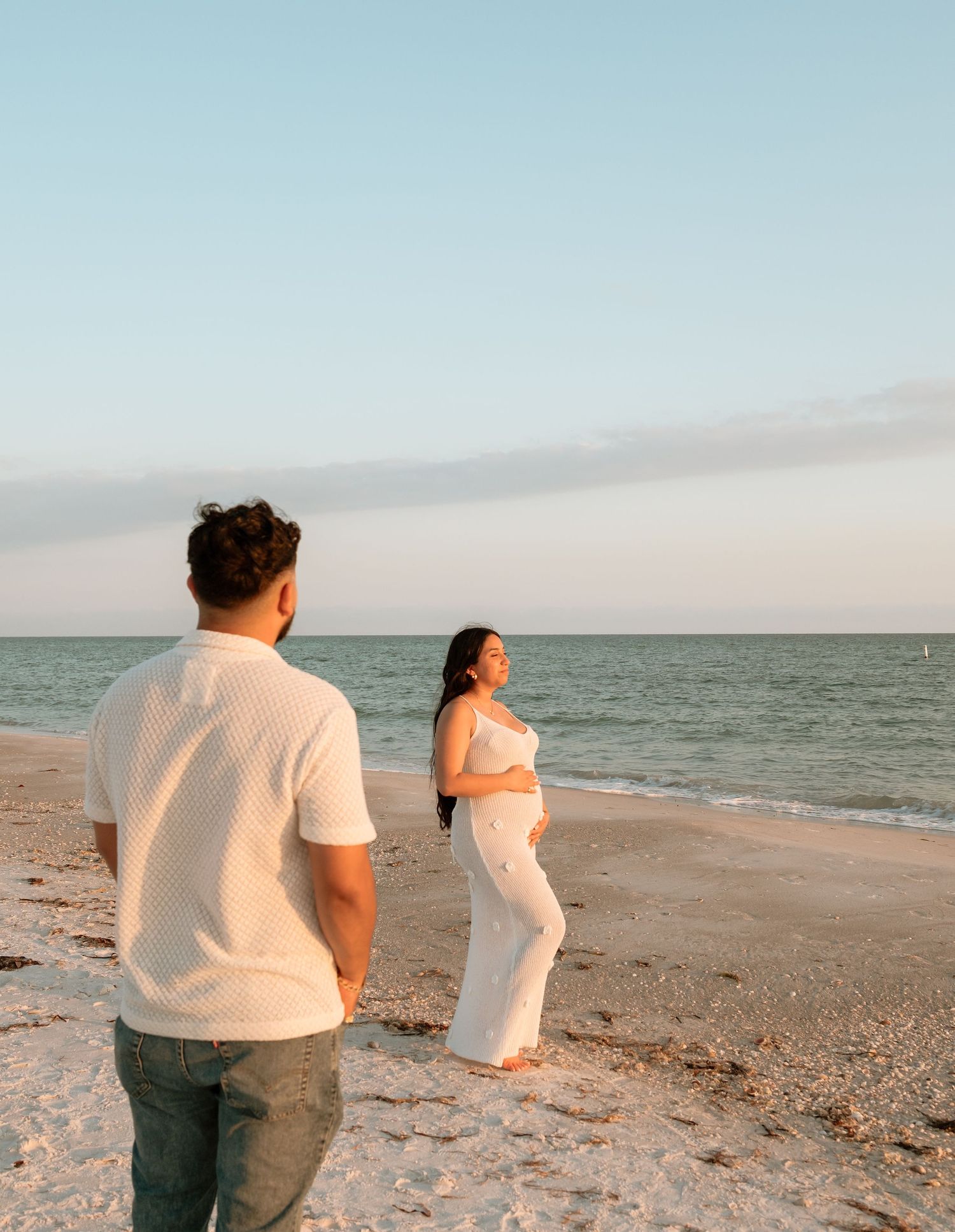 Couple on a sandy beach at sunset, woman in white outfit cradling baby bump while man in white shirt watches the ocean.