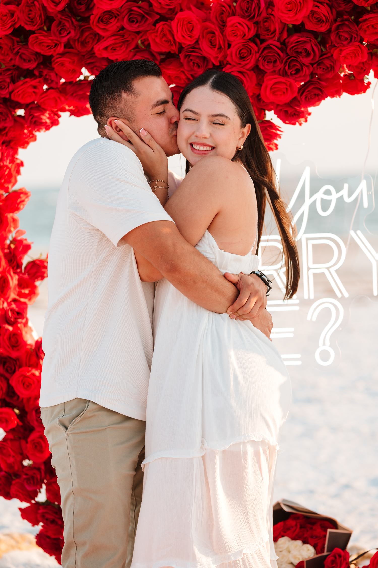 Couple embracing under a red rose arch on a beach, with a romantic marriage proposal moment captured outdoors.