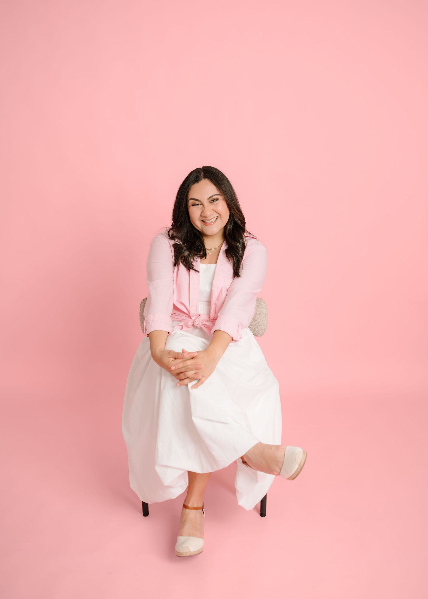 Smiling woman in white dress and pink cardigan sitting on chair against pink background.