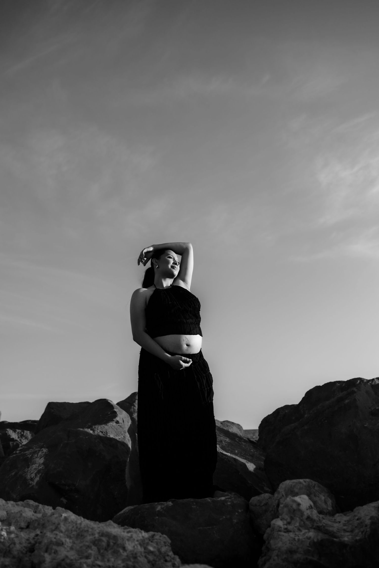 A dramatic black and white photo of a person in a black dress posing on rocky terrain against a cloudy sky.