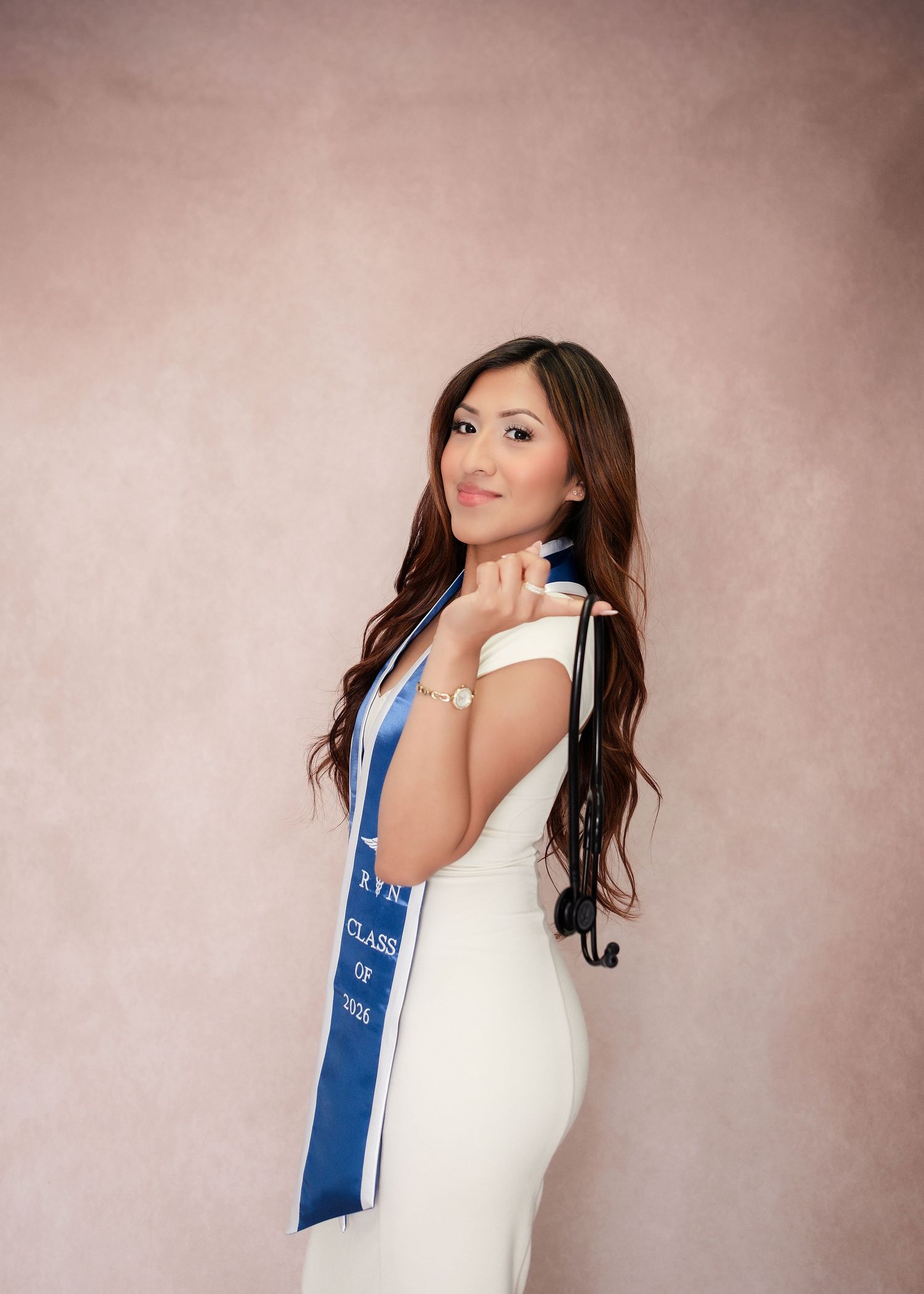 Young woman in white dress wearing blue graduation sash poses for senior portrait photo.