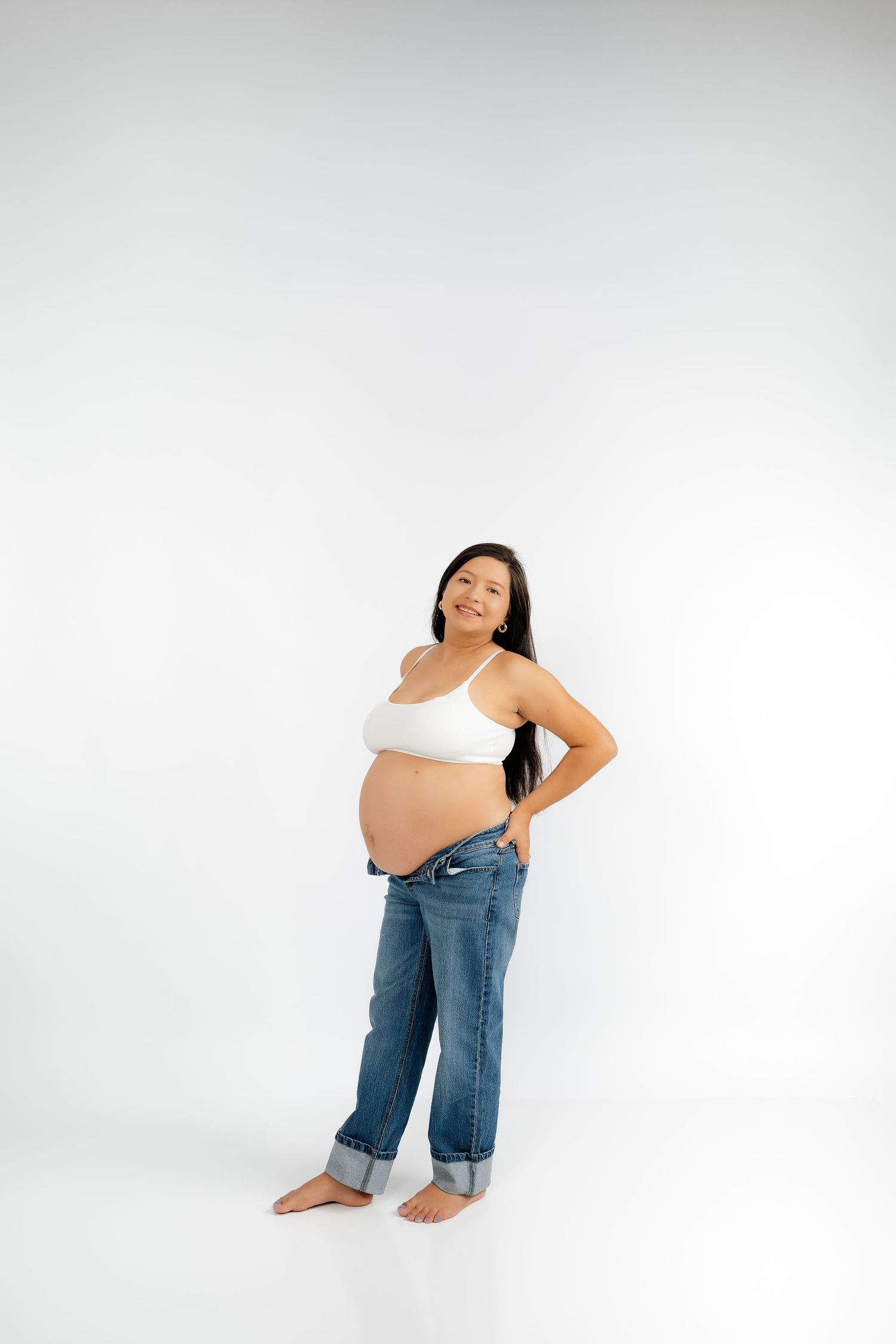 Smiling pregnant woman in white crop top and rolled jeans posing barefoot against white background.