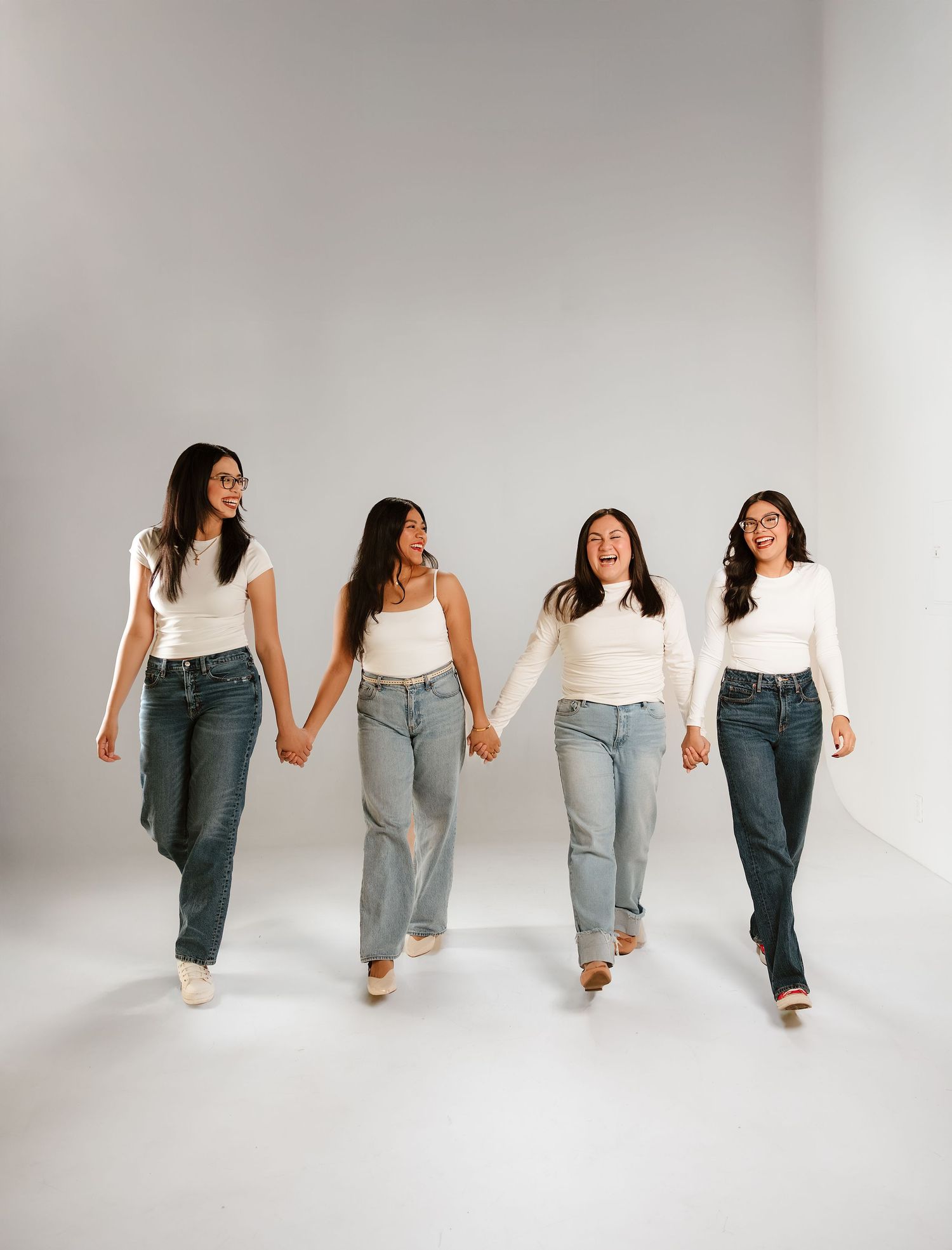 Four young women in white tops and light jeans walk hand-in-hand, smiling and laughing in a bright studio setting.
