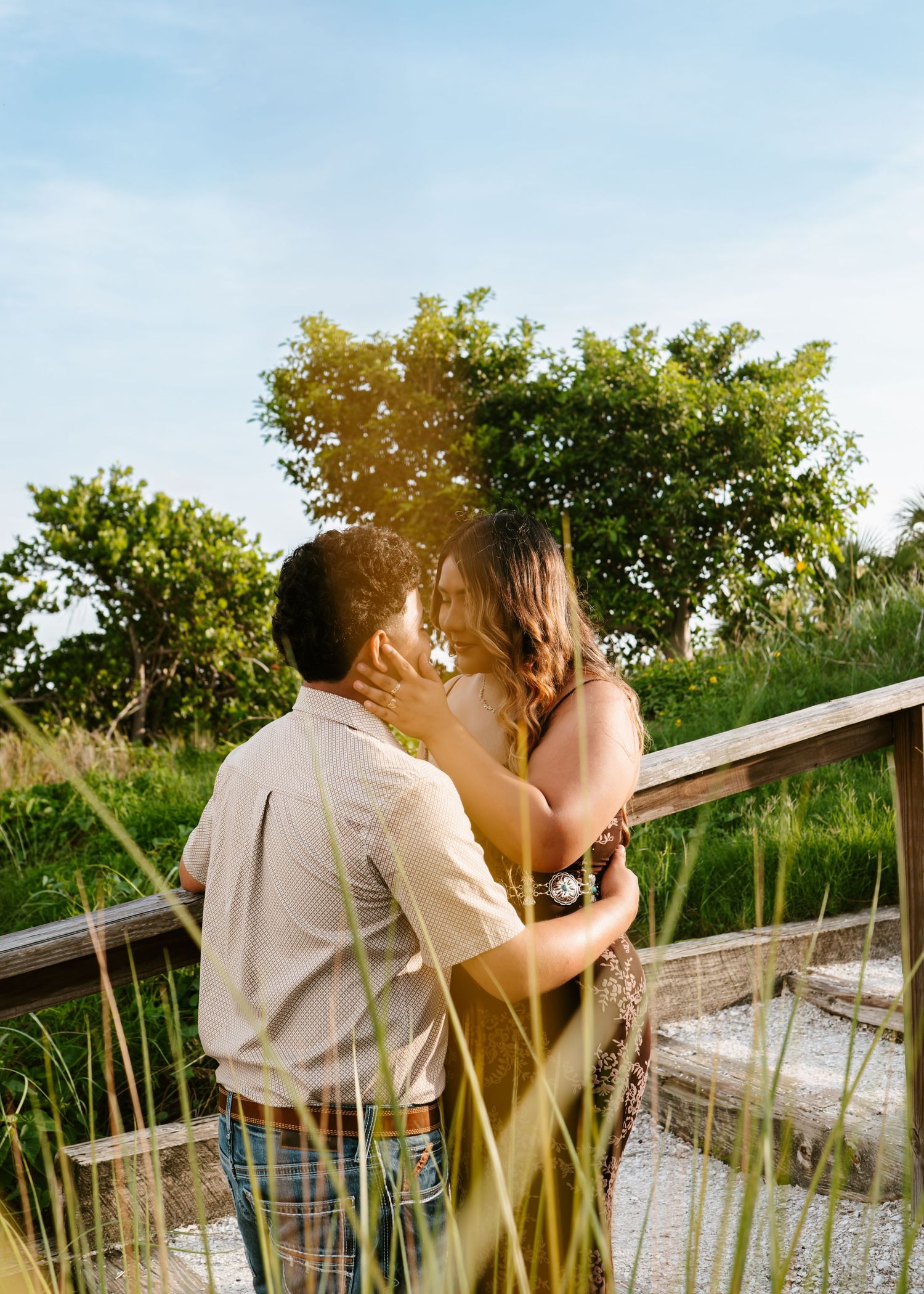 Couple sharing a romantic kiss on a wooden boardwalk surrounded by lush greenery in golden sunlight.