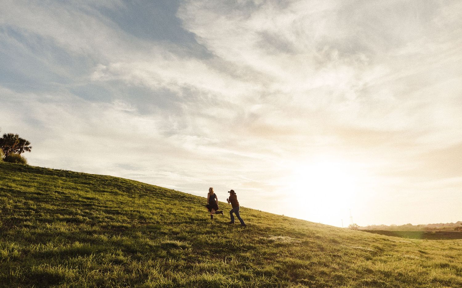 Two people walking up a lush green hillside at sunset under a dramatic cloudy sky.