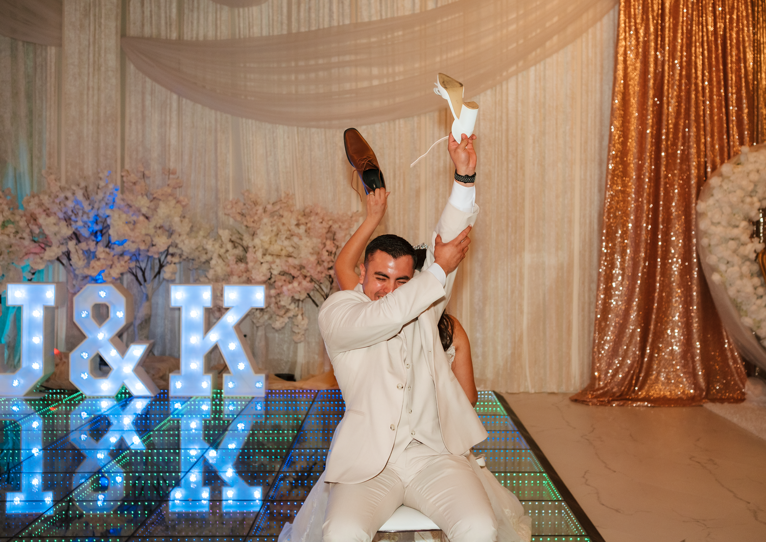 Groom in white suit dancing energetically at wedding reception with illuminated J&K monogram letters.