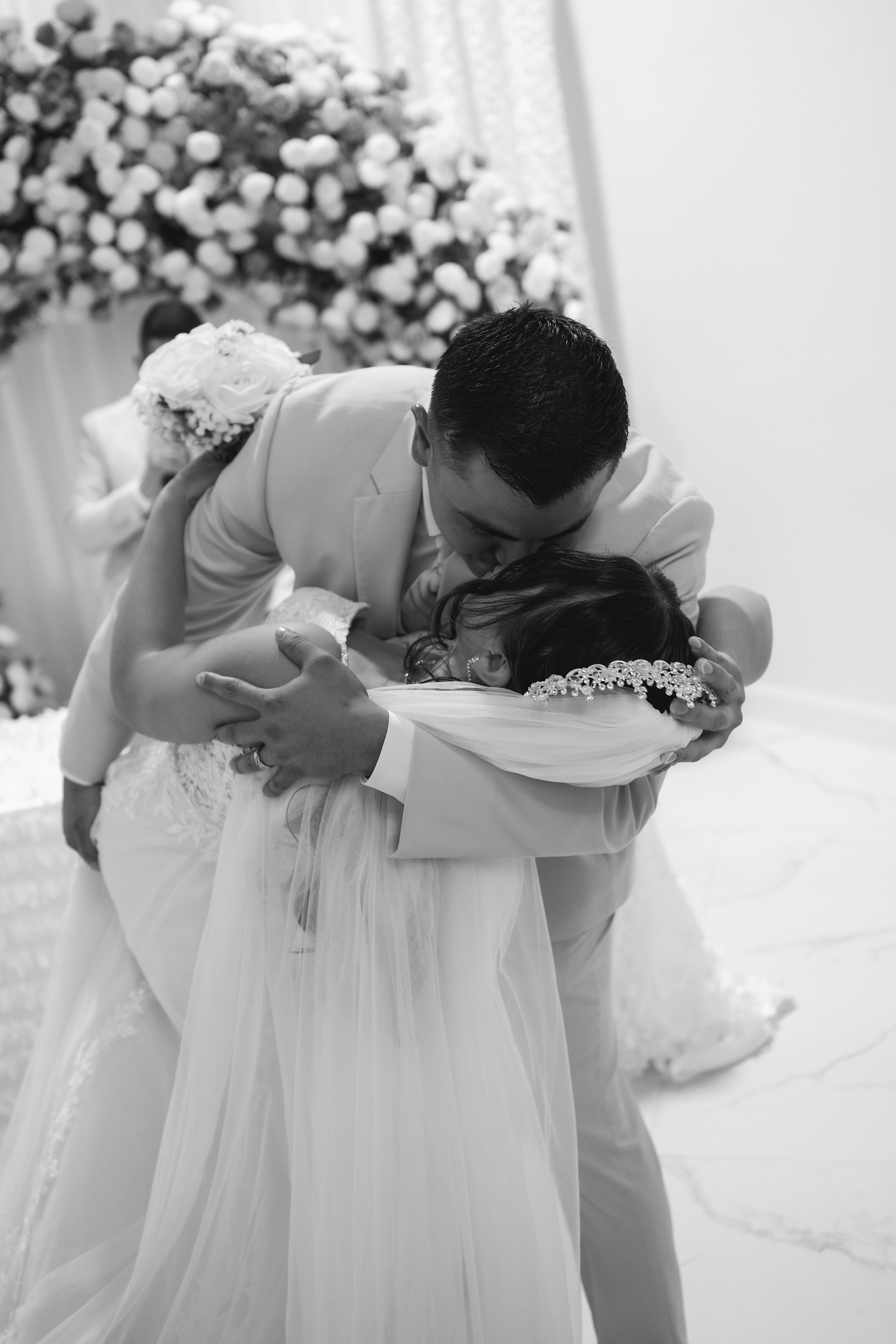 Black and white photo of bride and groom sharing an emotional embrace at the wedding altar with floral backdrop.