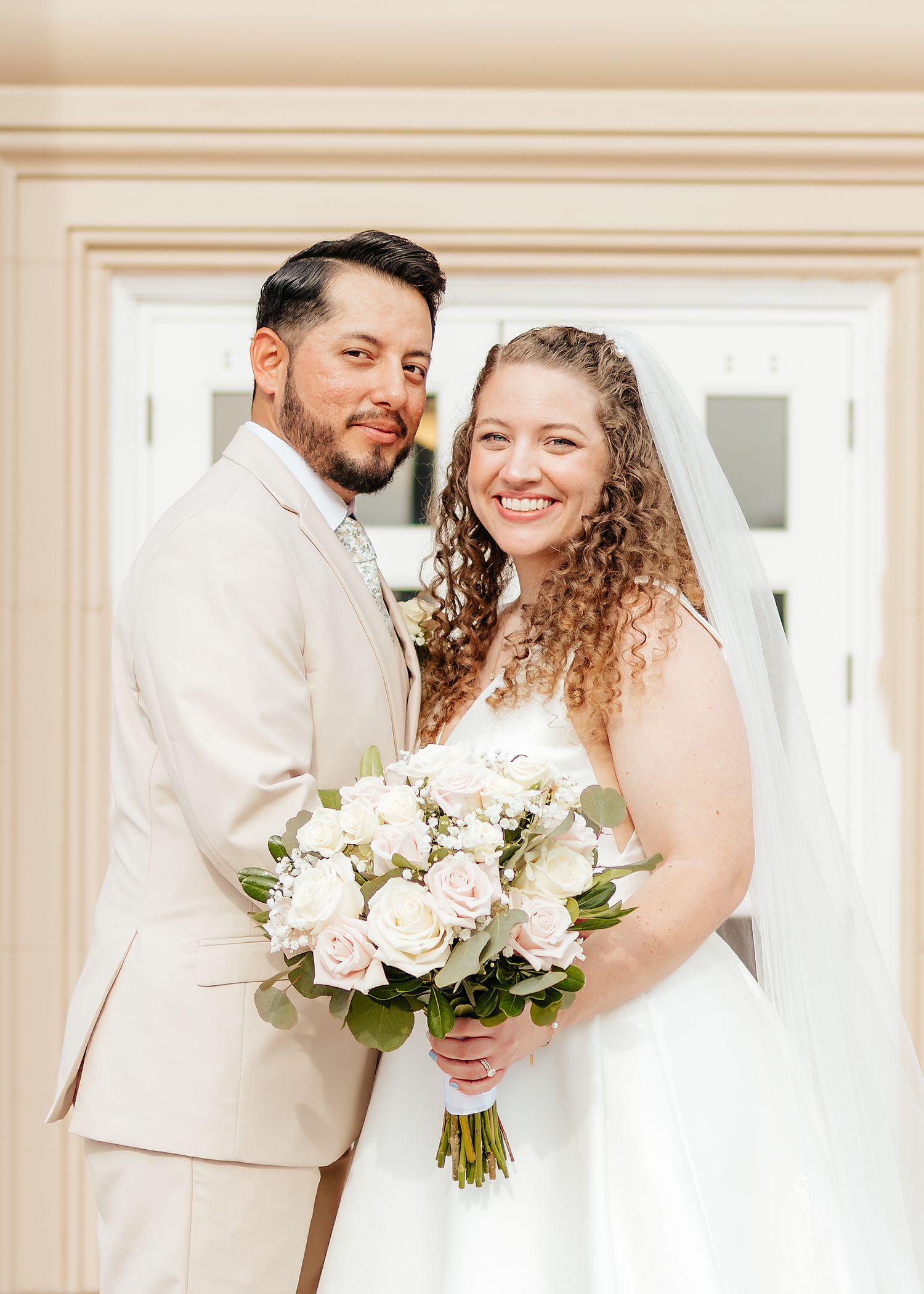 Smiling bride with curly hair holds white rose bouquet beside groom in tan suit for wedding portrait.