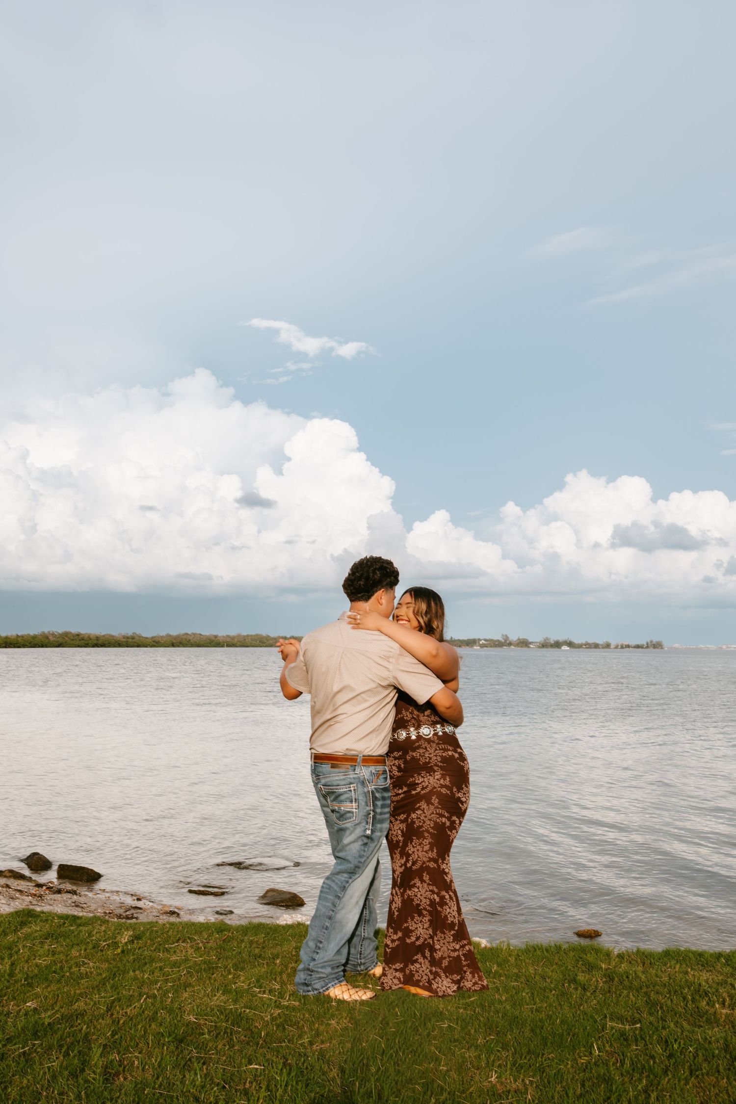 Couple embracing and kissing by a lakeside on a grassy shore with dramatic clouds and calm water in the background.