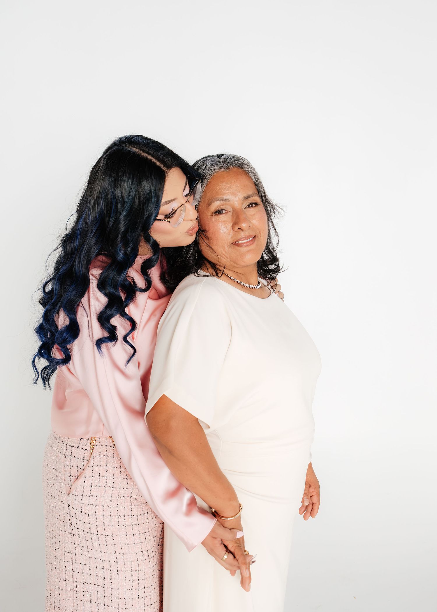 Two women in pink and white outfits pose together, one kissing the other on the cheek against a white background.
