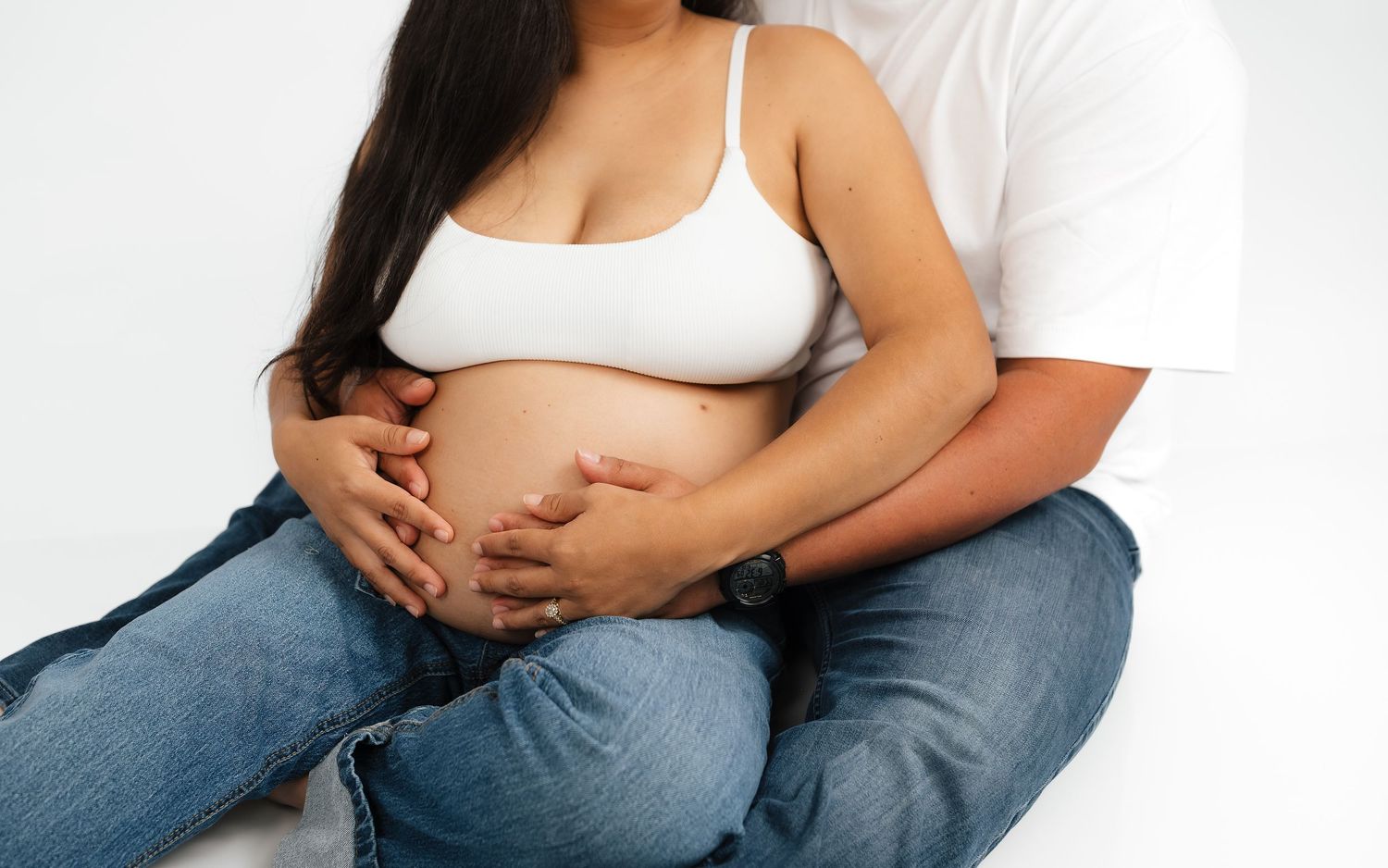 Pregnant woman in white bra and jeans sitting with partner's hands on baby bump against white background.