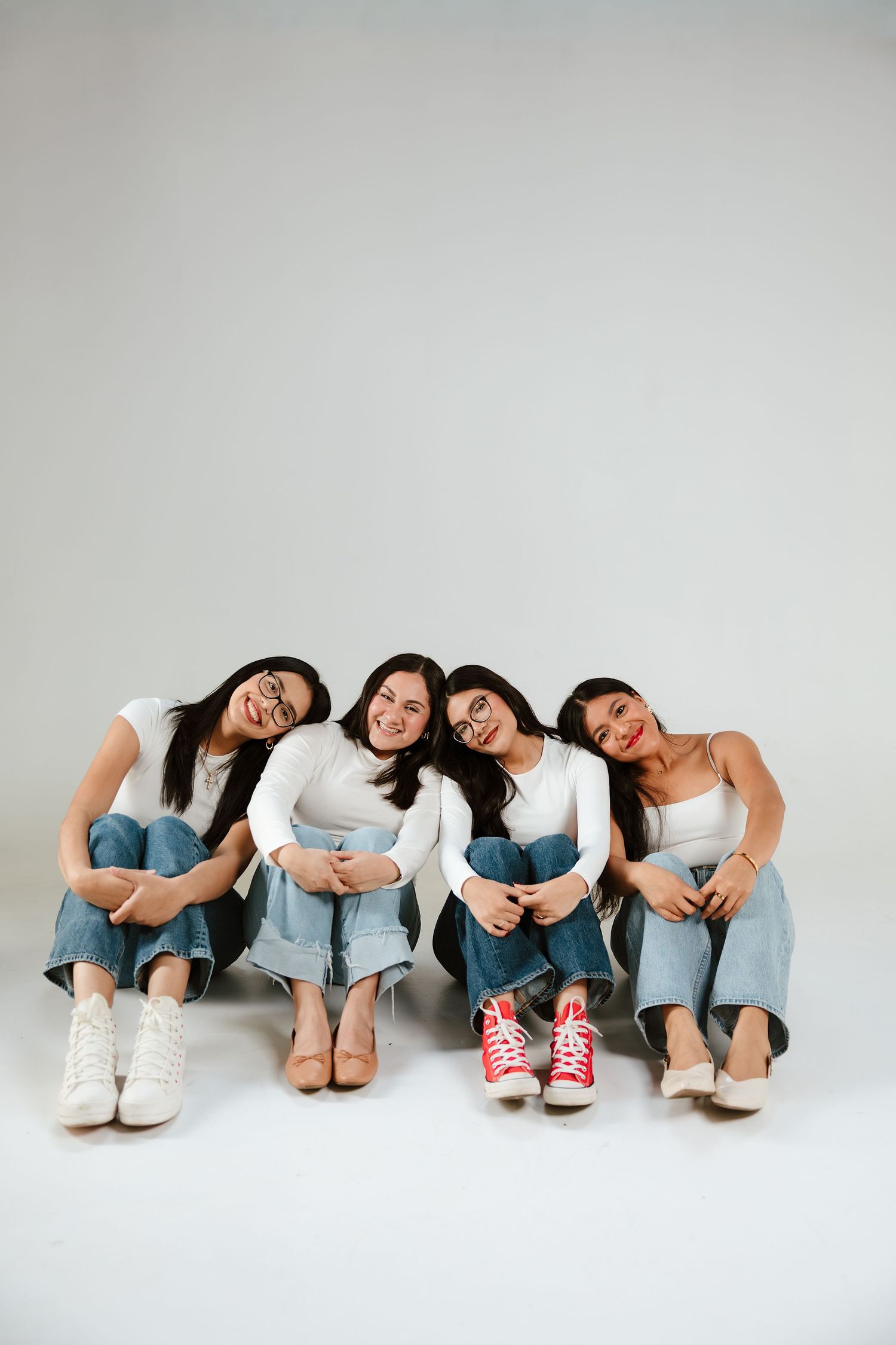 Four young women in white tops and jeans sitting together on floor, smiling and leaning on each other.