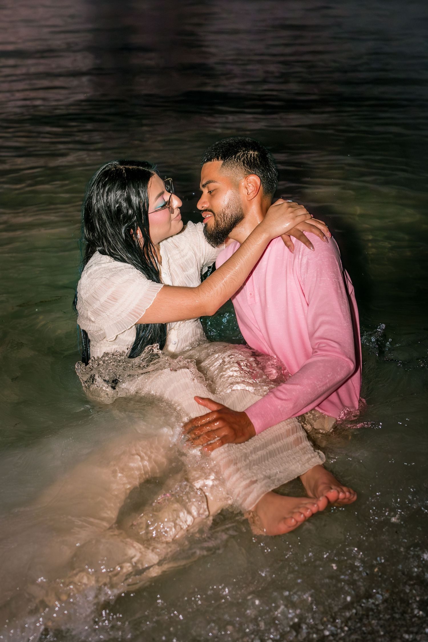 Couple sitting together in ocean waves at night, woman in white dress embracing man in pink sweater on beach.
