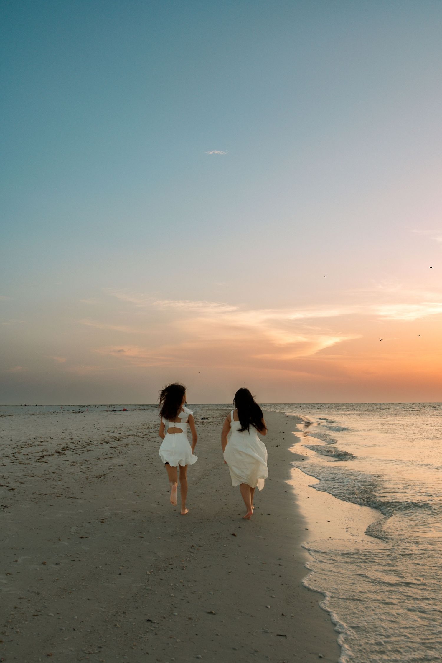 Two people in white dresses walk along a serene beach at sunset with waves gently rolling onto the shore.