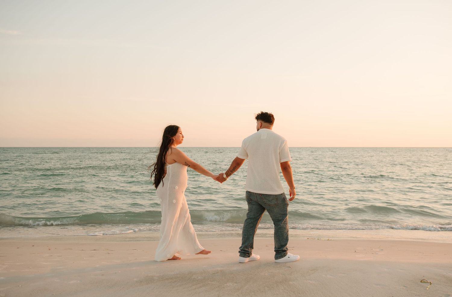 Two people walk hand in hand along a serene beach at sunset with ocean waves rolling in.