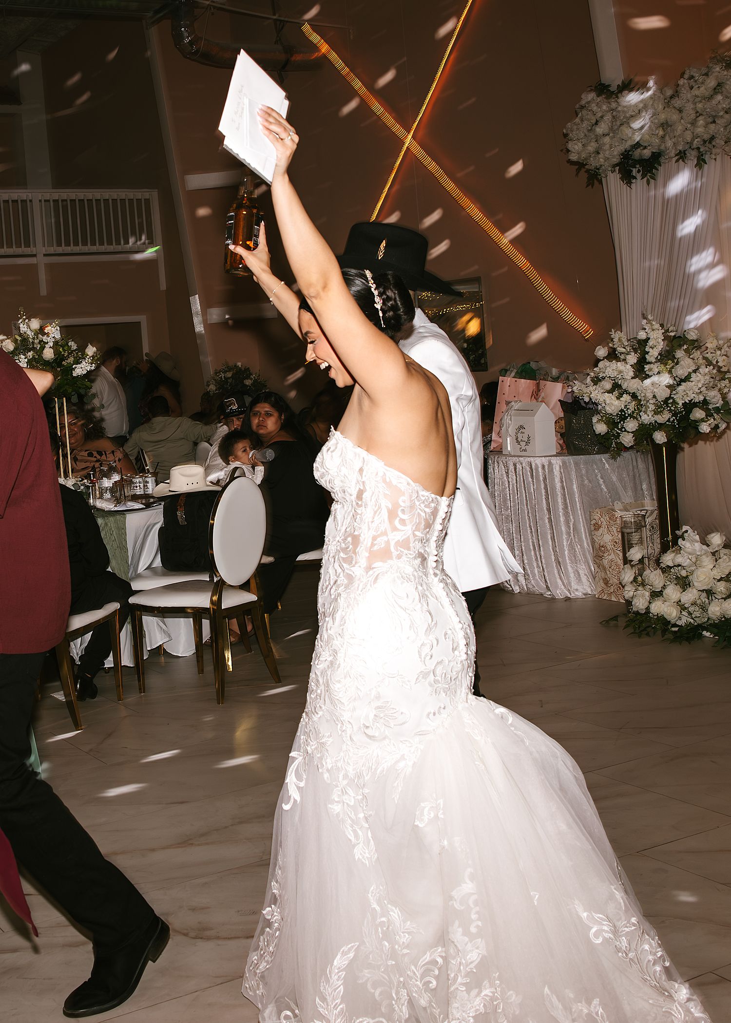 Bride in lace mermaid wedding gown raises arm joyfully on dance floor at elegant wedding reception.