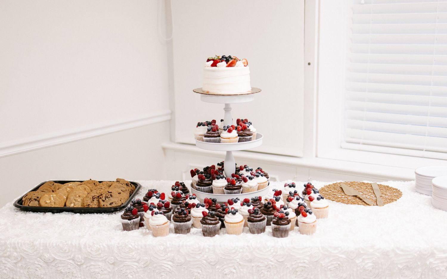 Elegant wedding dessert table with tiered cake, cupcakes, and cookies on white linen.
