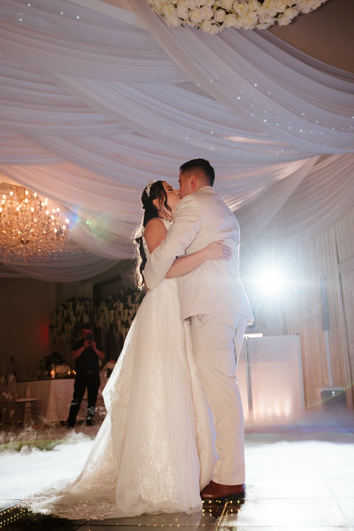 Bride and groom sharing first dance kiss under white draped ceiling with chandelier and dramatic stage lighting.