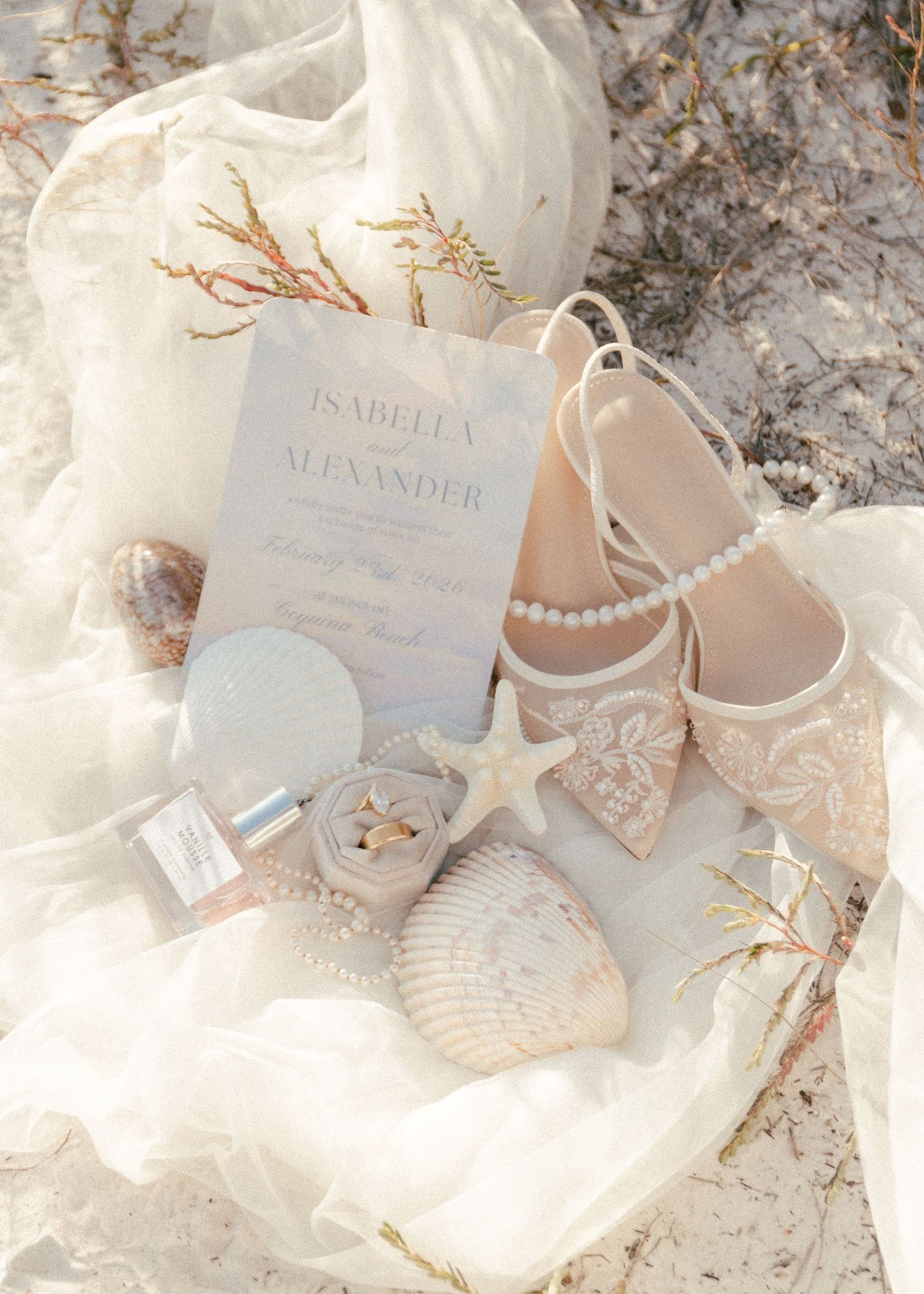 Beach wedding flat lay with pearl shoes, starfish, shells, rings, and Isabella & Alexander invitation on white fabric.