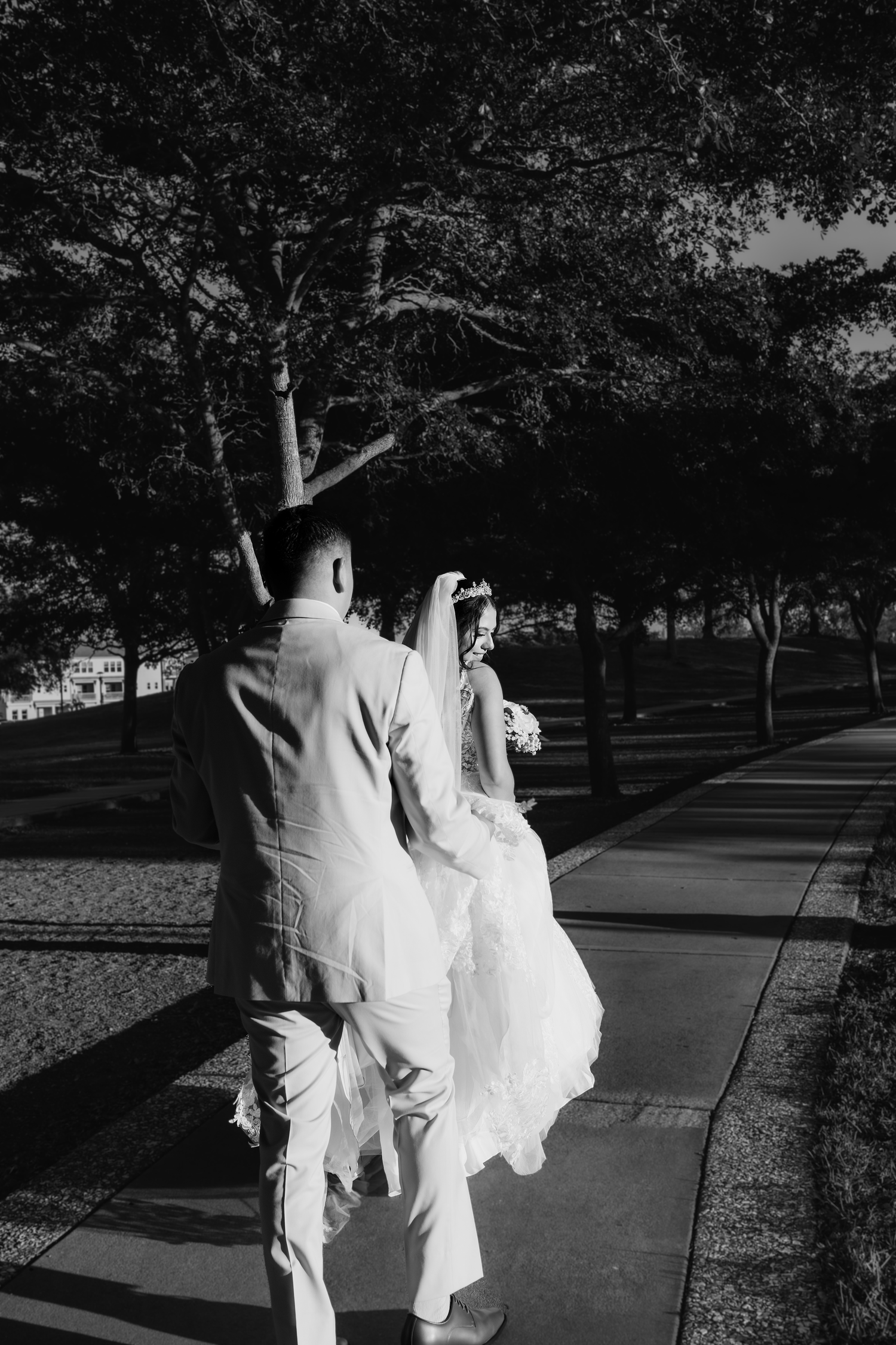 Black and white photo of bride and groom walking hand in hand along tree-lined path after wedding.
