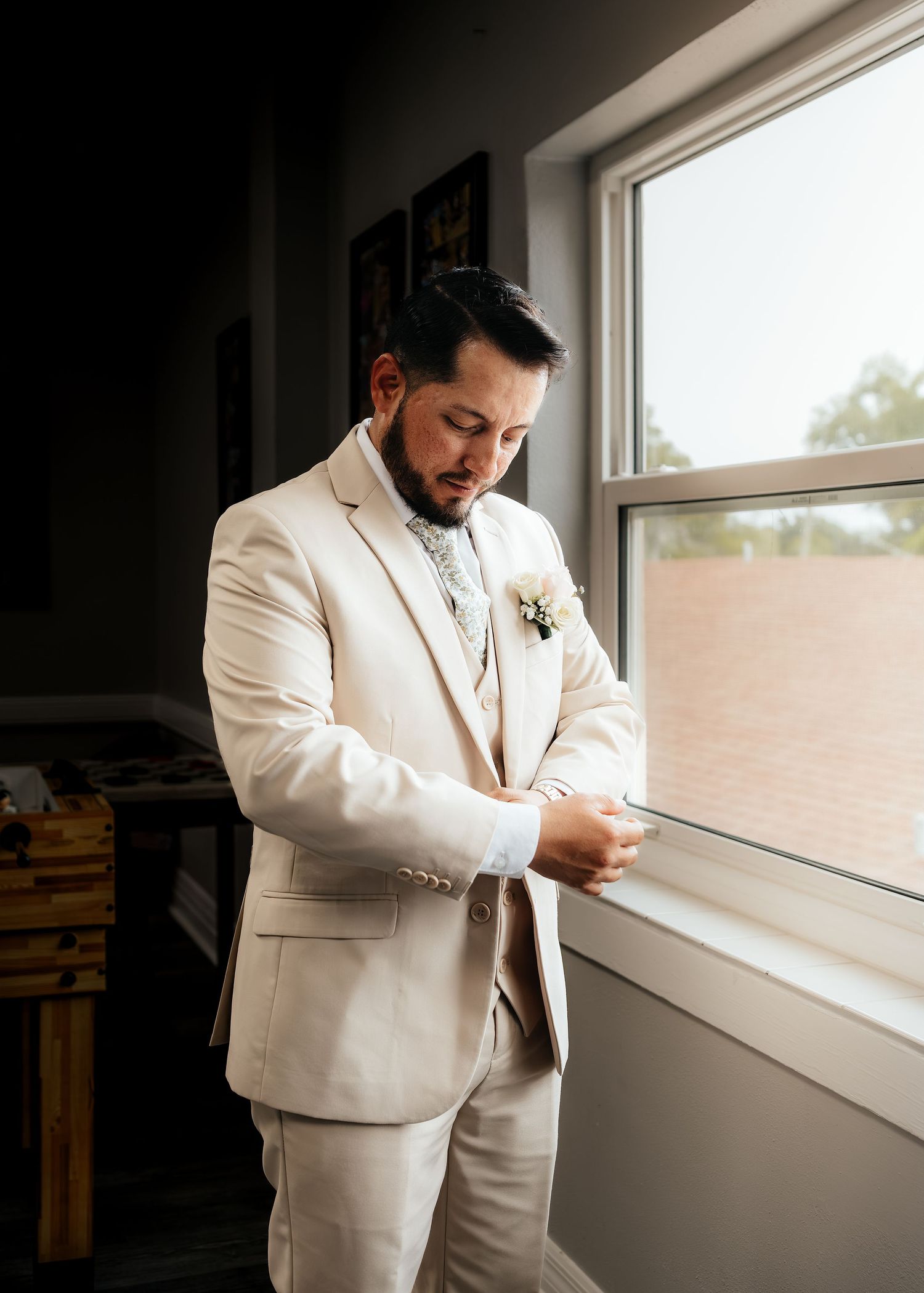Groom in a beige three-piece suit adjusting his cufflinks by a window with a floral boutonniere.