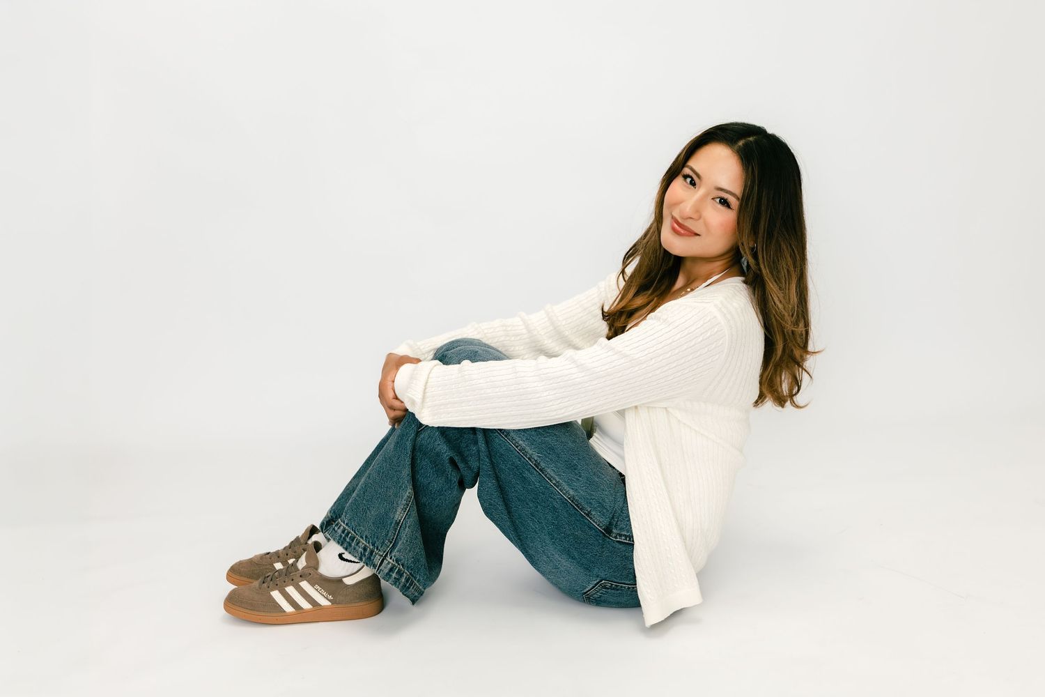 Young woman sitting on floor in white sweater, jeans, and Adidas sneakers against white background.