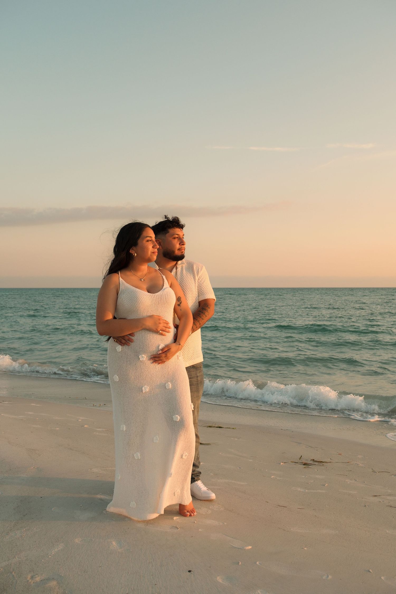 Pregnant couple embracing on a sandy beach at sunset, woman in white dress, ocean waves in background.