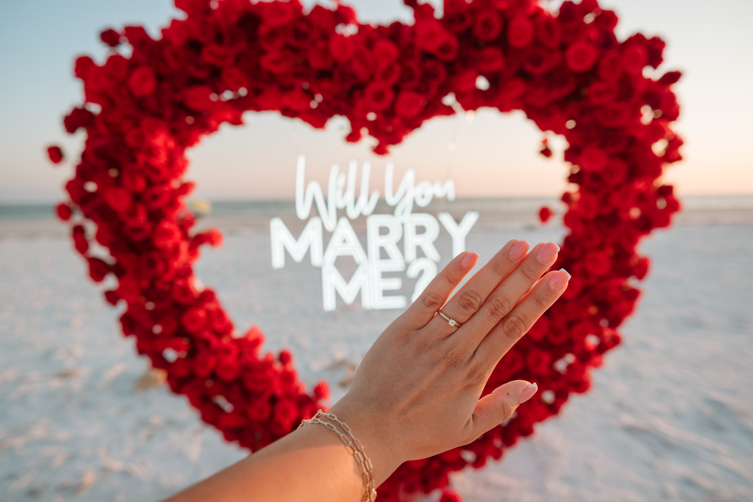 Hand with engagement ring held up before a red rose heart shape with Will You Marry Me text on a beach.