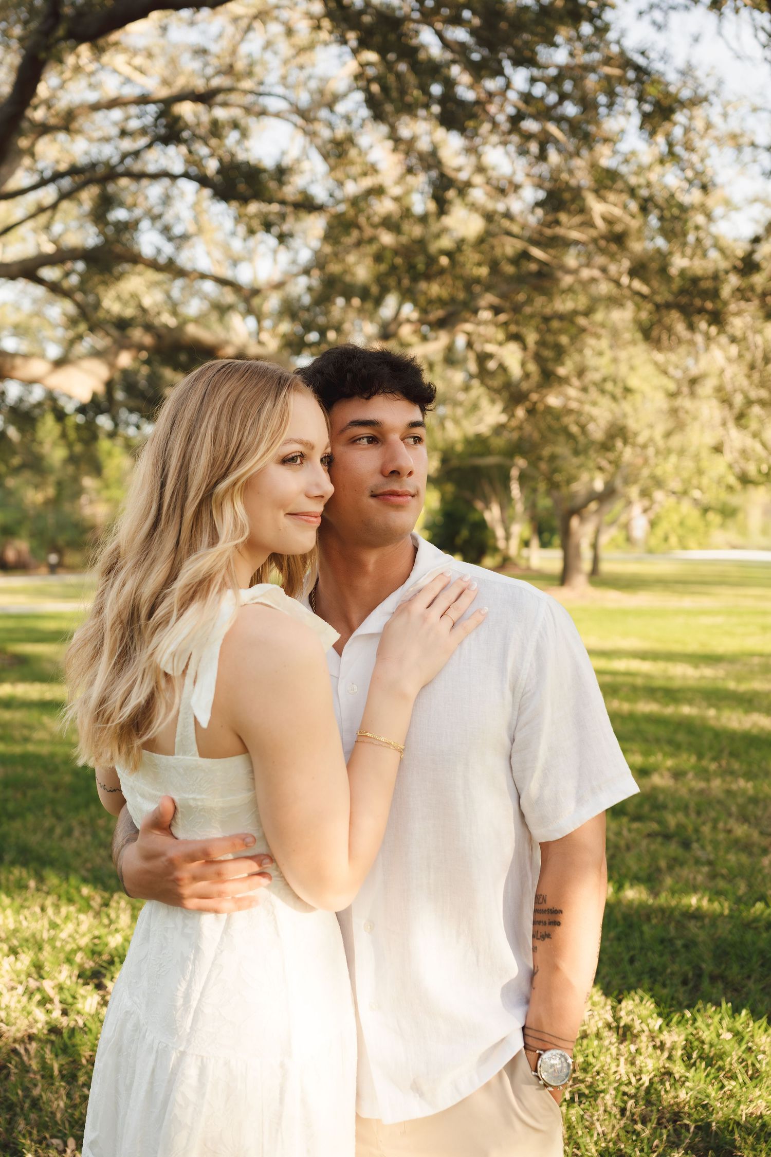 Couple embracing in a sunny park, woman in white dress with blonde hair, man in white shirt smiling outdoors.