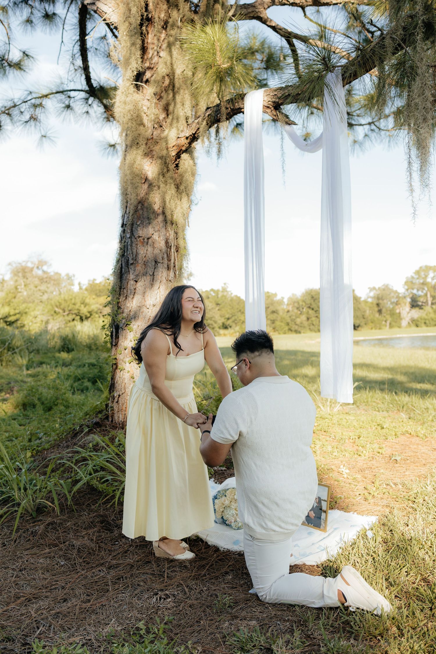 Man kneeling on one knee proposing to a smiling woman in a yellow dress under a decorated tree arch outdoors.