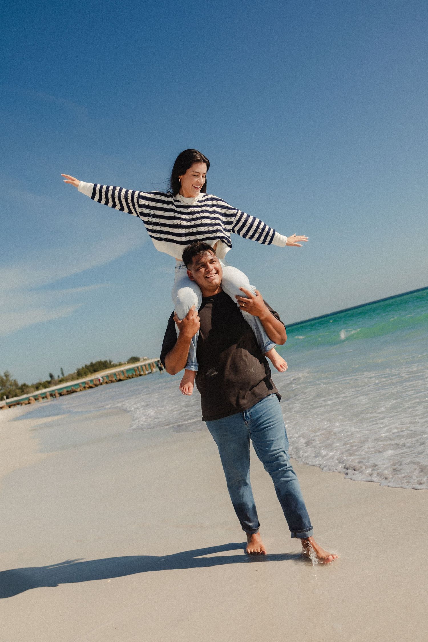 A joyful couple on a sunny beach, man carrying woman on his shoulders as she spreads her arms wide like airplane wings.