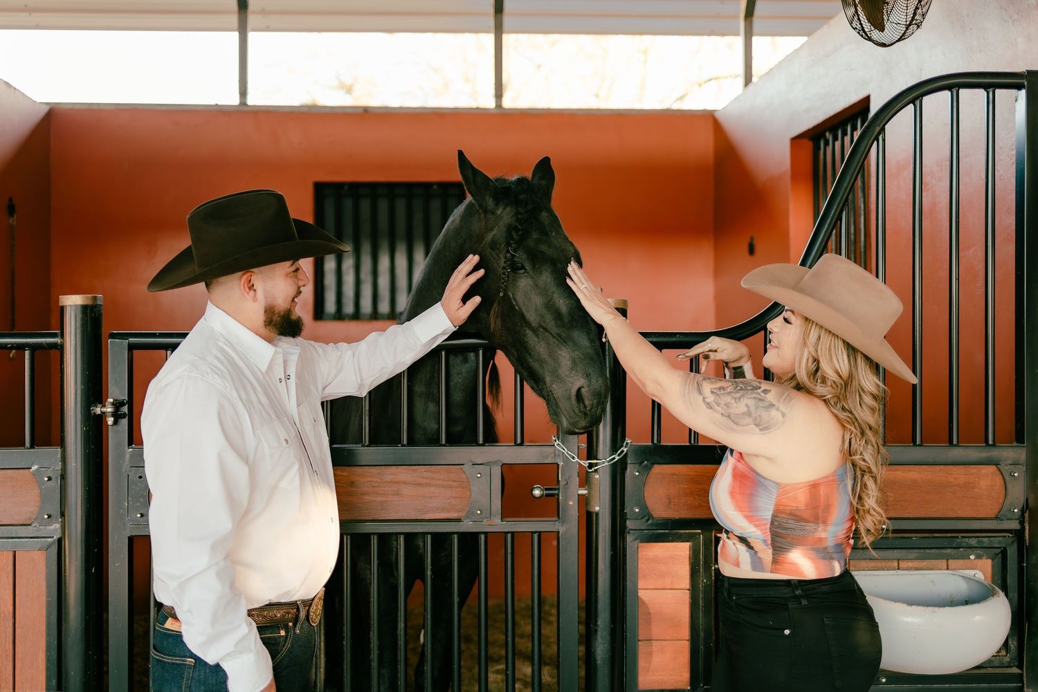 A couple in cowboy hats petting a black horse in a rustic stable with red walls.