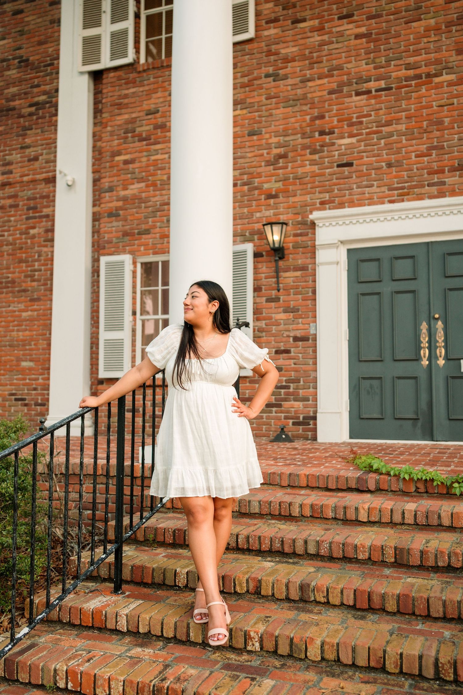 Young woman in white dress posing on brick steps outside a classic red brick building with green doors.