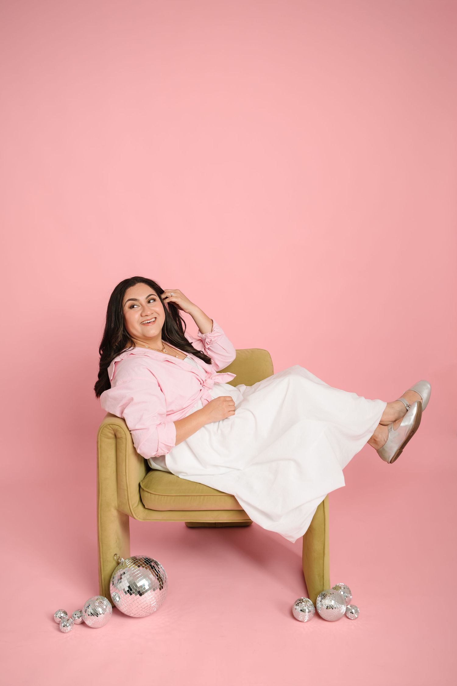 Woman in pink blouse and white skirt relaxing in olive chair surrounded by silver disco balls on pink background.