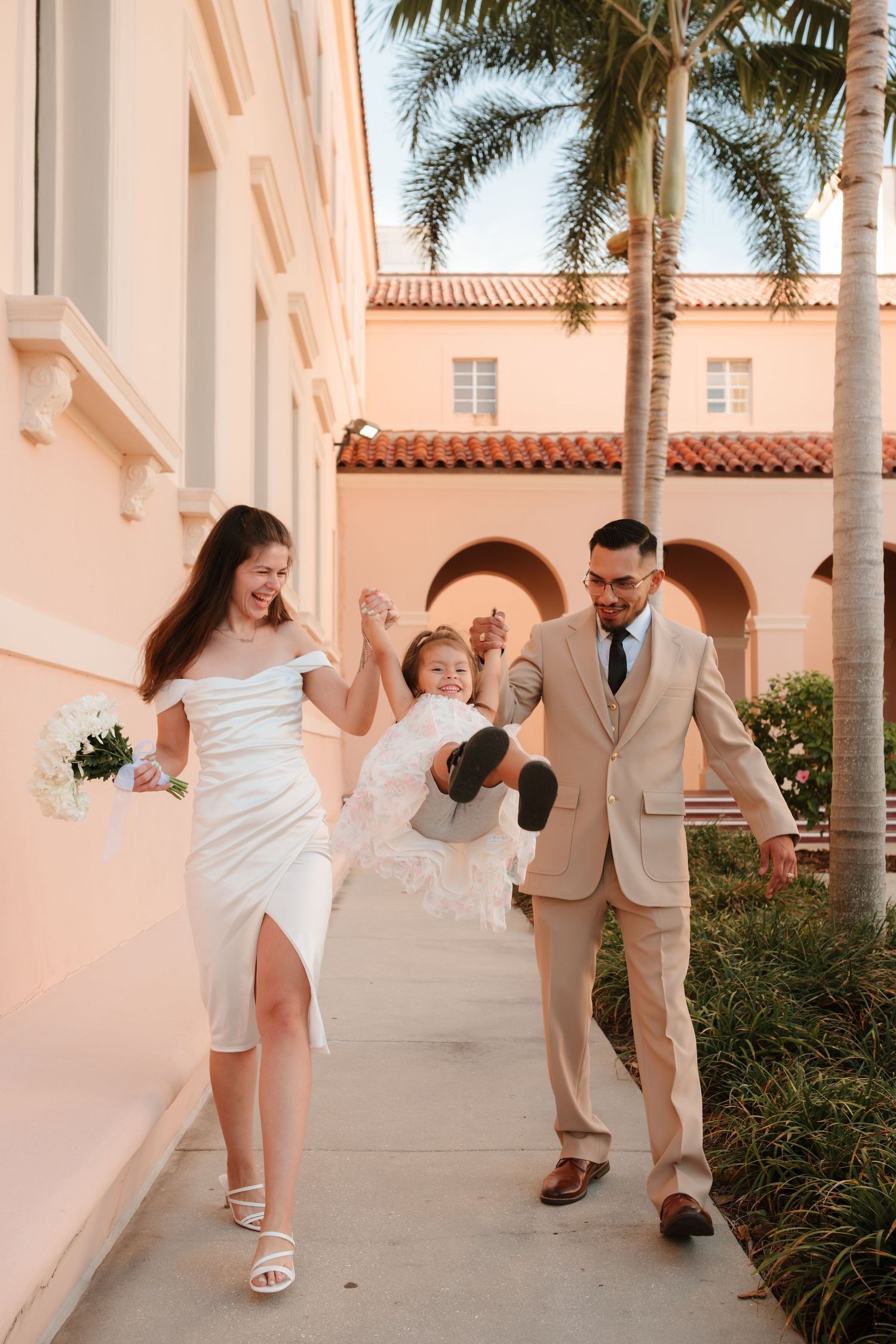 Bride and groom in wedding attire swing their young daughter between them outside a Spanish-style building with palm trees.