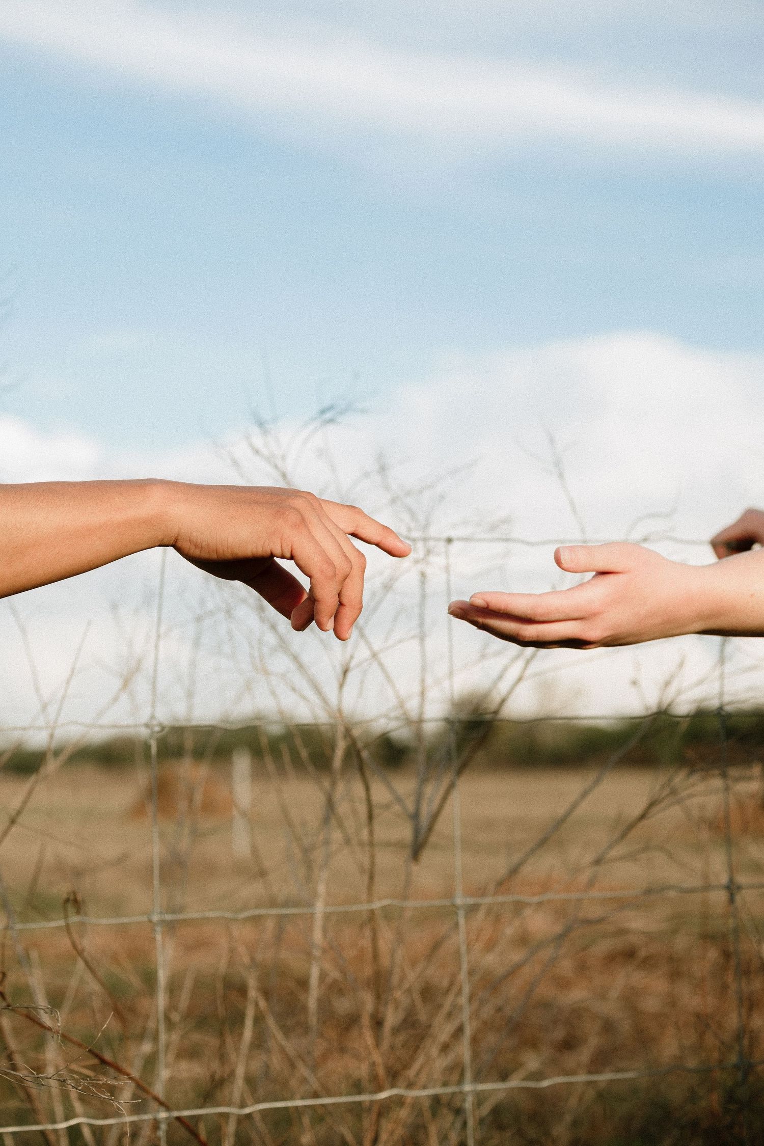 Two hands reaching toward each other outdoors, evoking connection and trust against a rural field and cloudy sky backdrop.