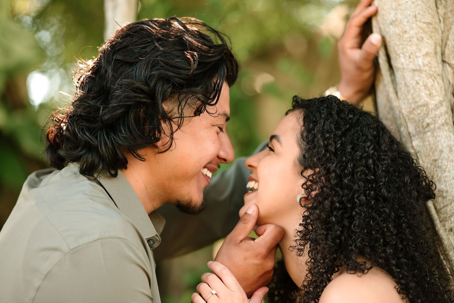 A smiling couple gazes at each other romantically near a tree in an outdoor setting.